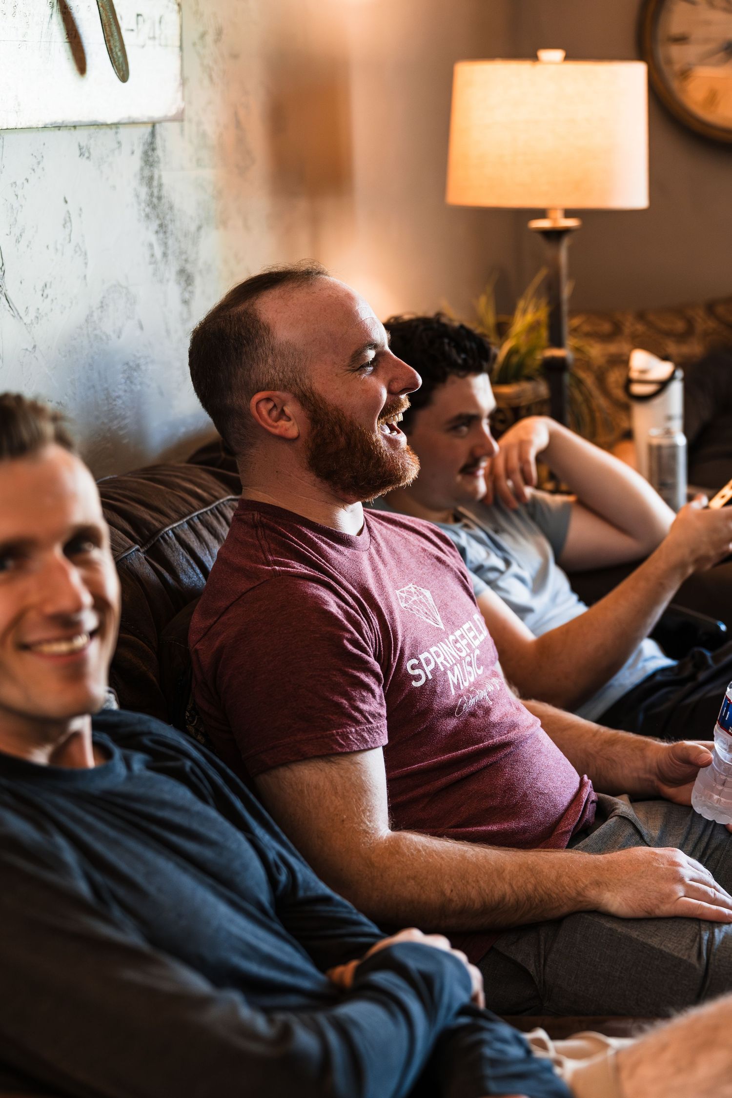 Three men laughing and relaxing together on a couch in a warmly lit living room.