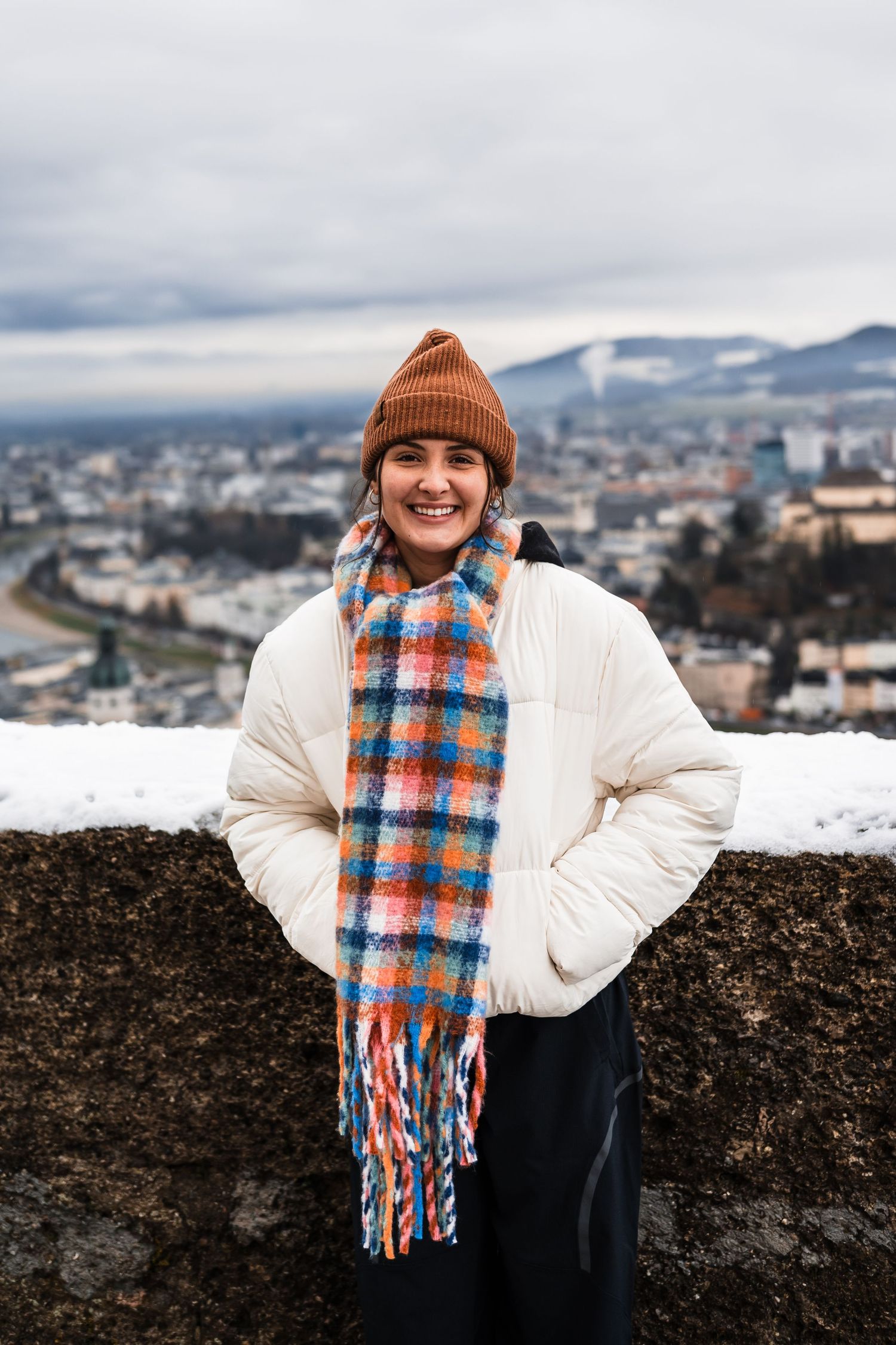 Person in white winter coat and orange beanie with plaid scarf poses against snowy cityscape backdrop.