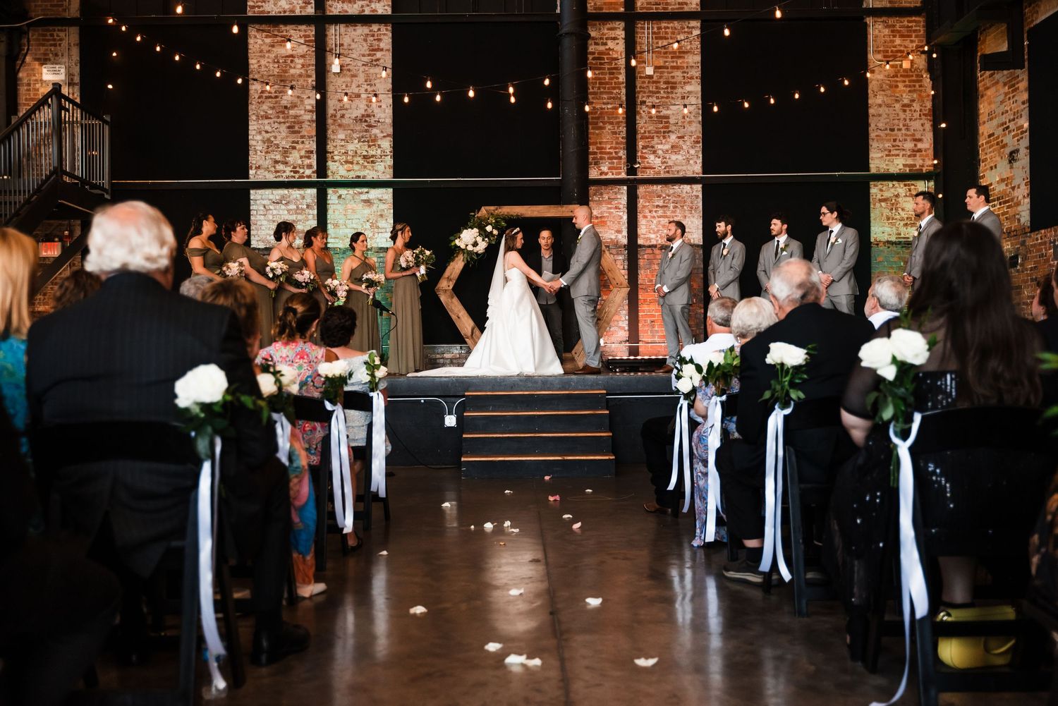 A wedding ceremony in a rustic brick venue with string lights overhead and white floral decorations along the aisle.