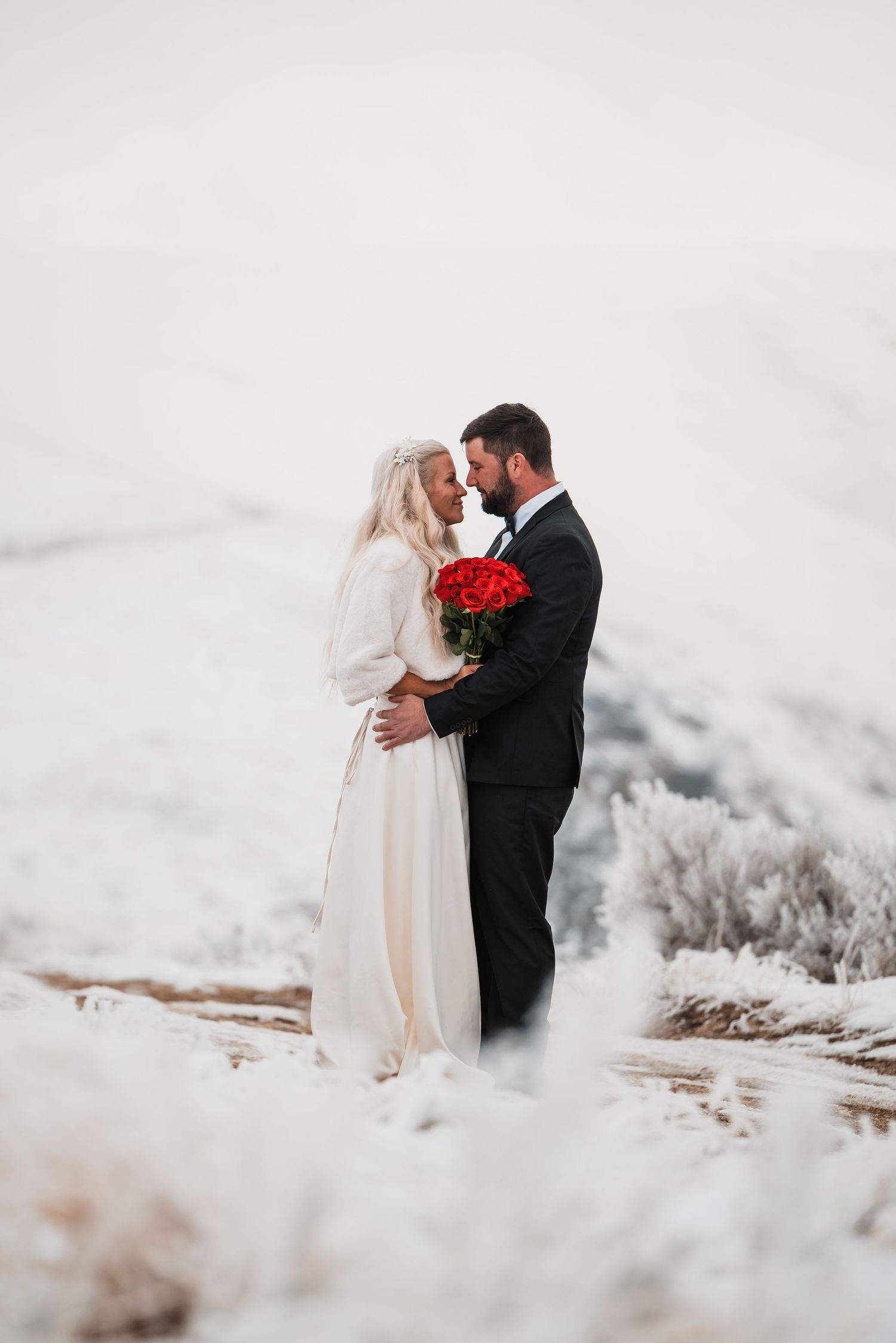 Romantic winter wedding portrait with red roses against snowy mountain backdrop.