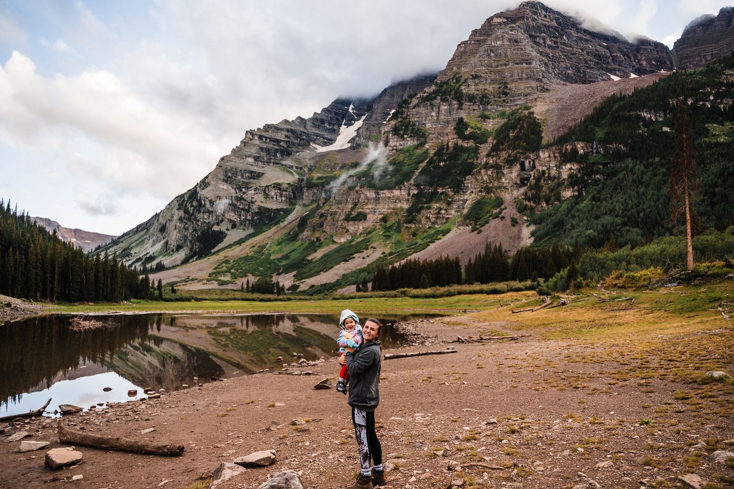 Person standing by mountain lake with dramatic peaks and reflective waters in background.