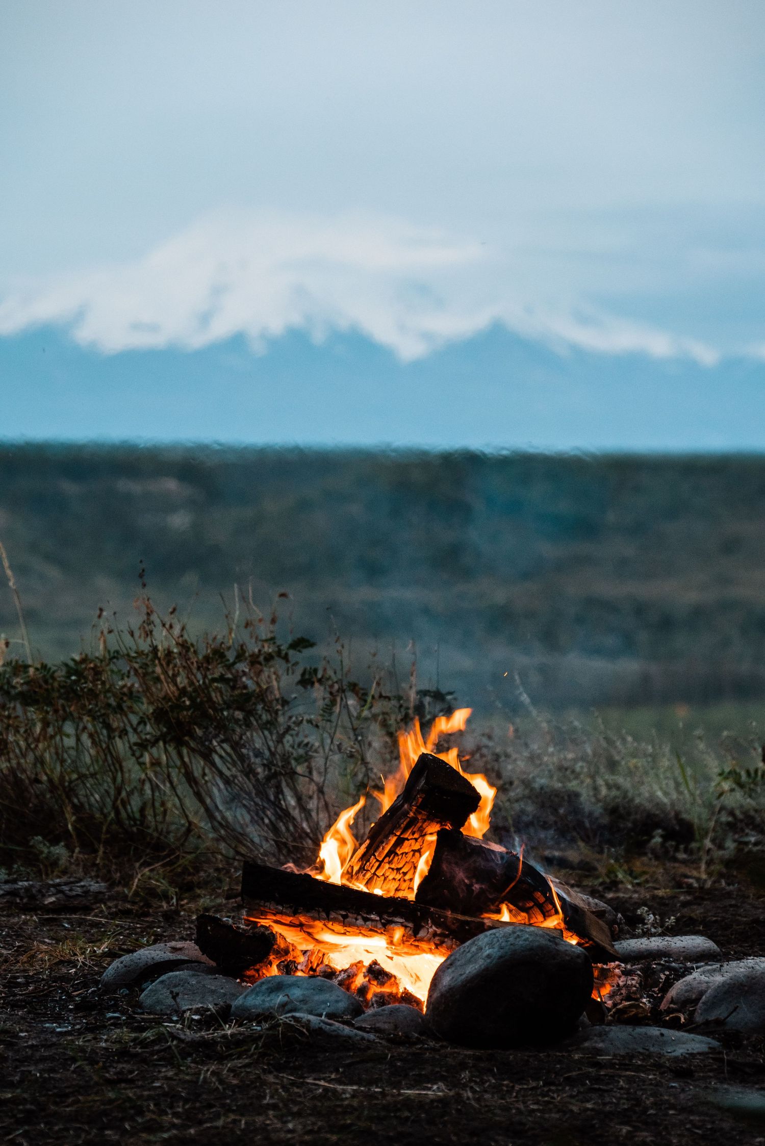 Campfire burns brightly against snowy mountain backdrop during blue hour.