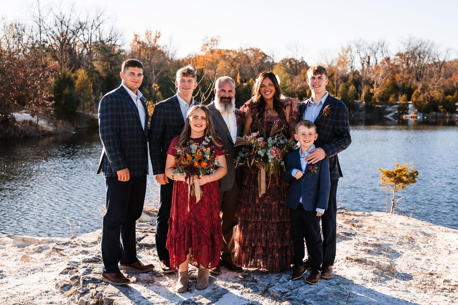 Family group poses together on rocky lakeside during autumn sunset wearing coordinated formal attire.