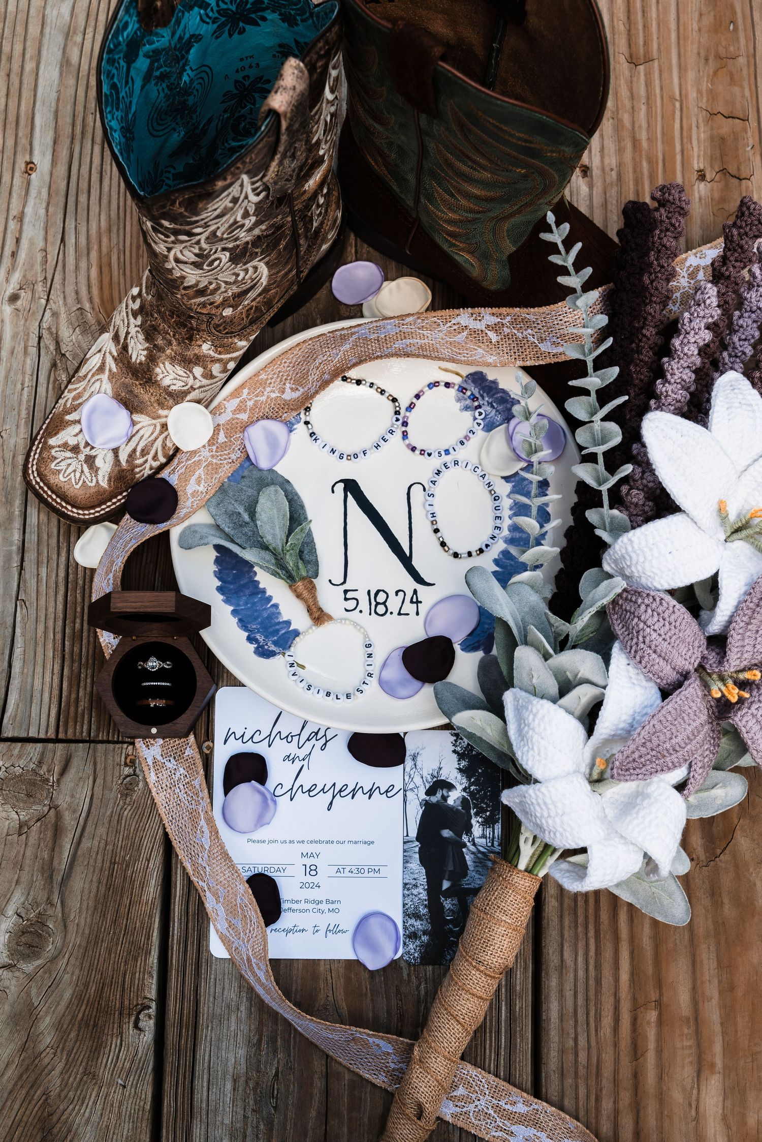 Rustic wedding details including cowboy boots, invitation, monogrammed plate, and floral arrangements on wooden planks.