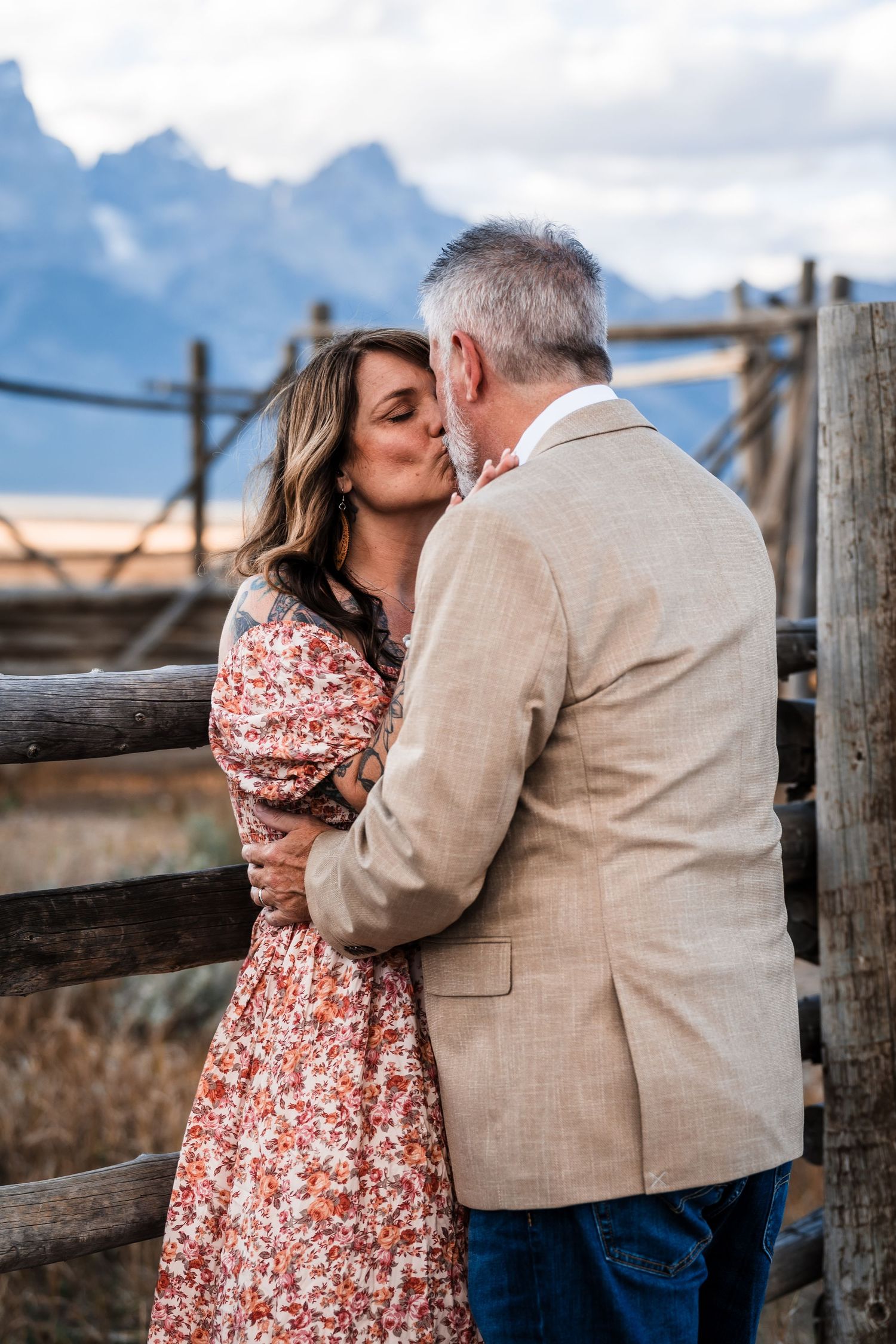 A couple embraces against a rustic wooden fence with mountains and cloudy skies in the background.