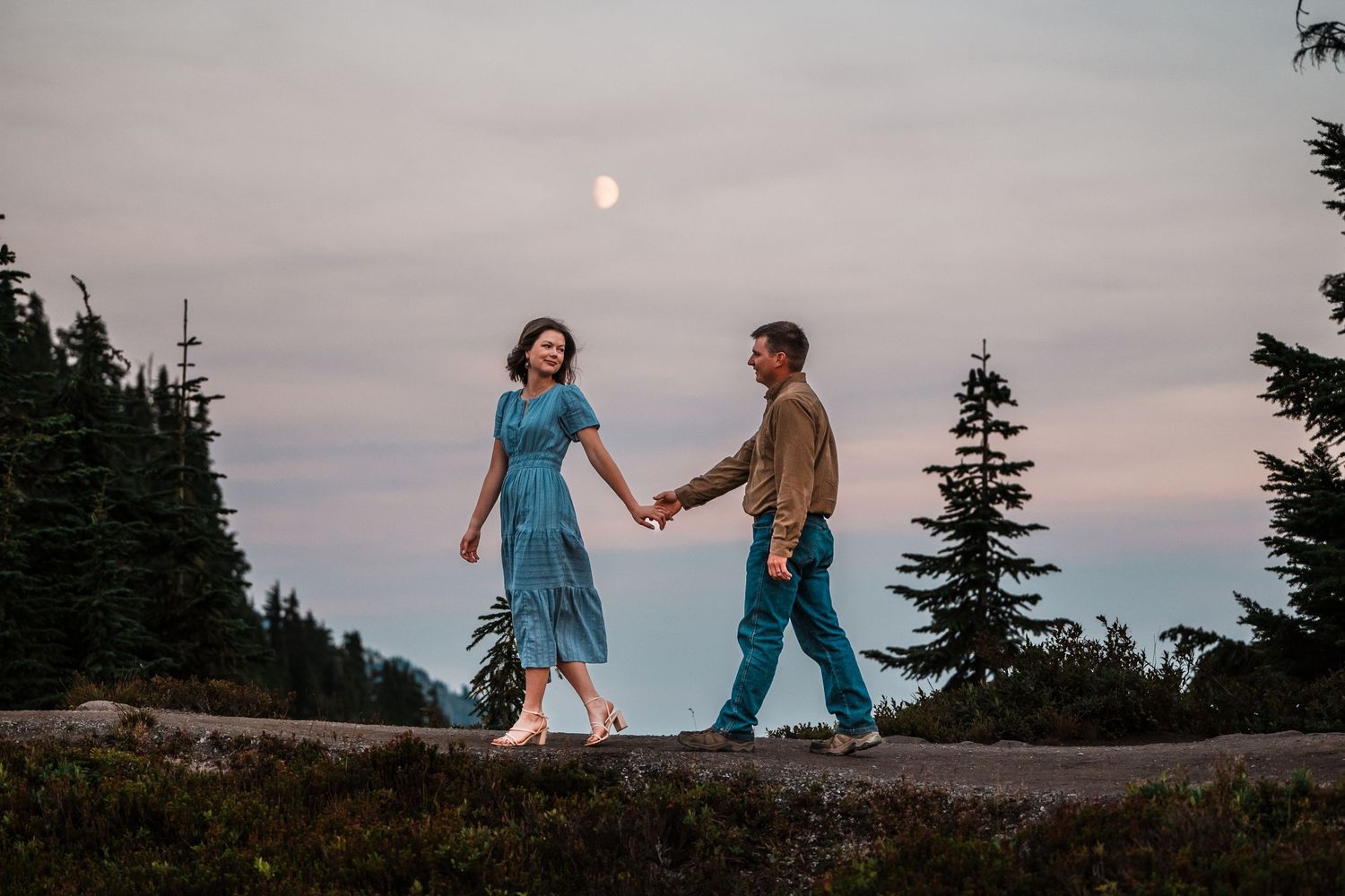 A couple dances together at sunset on a mountaintop with evergreen trees silhouetted against the dusky sky.