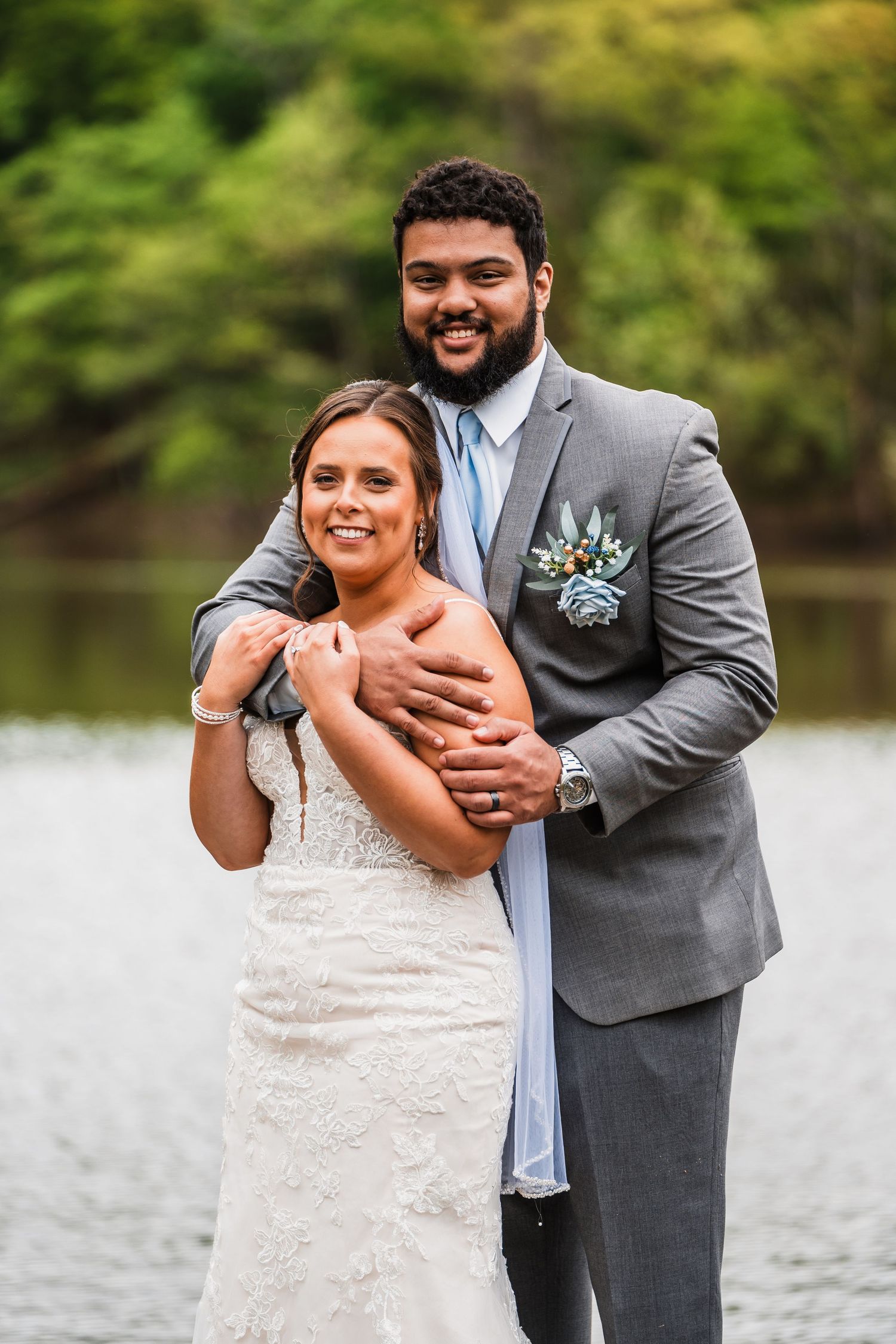 A couple poses for a wedding portrait by a lake wearing a lace gown and grey suit.