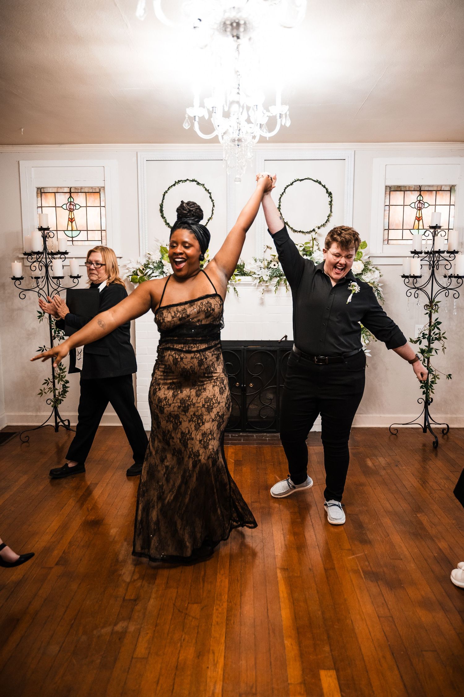 A couple dancing and celebrating with raised arms under a crystal chandelier at their wedding reception.