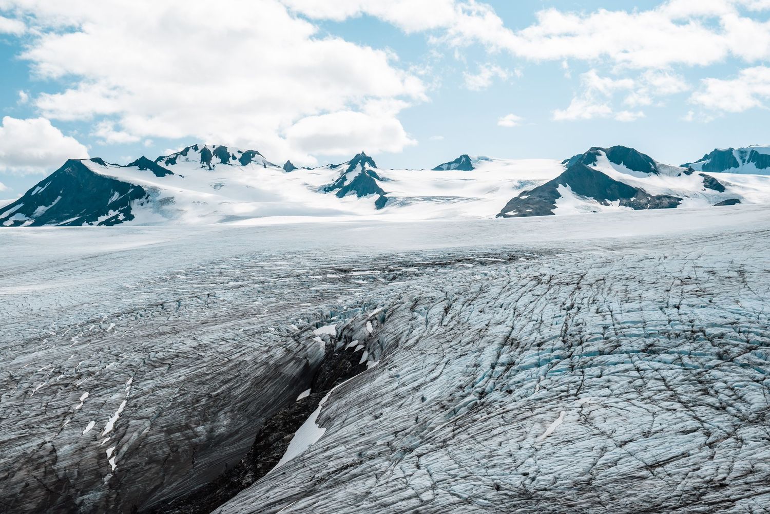 Snow-covered mountain peaks rise above a massive glacier with deep crevasses in Alaska's rugged wilderness.