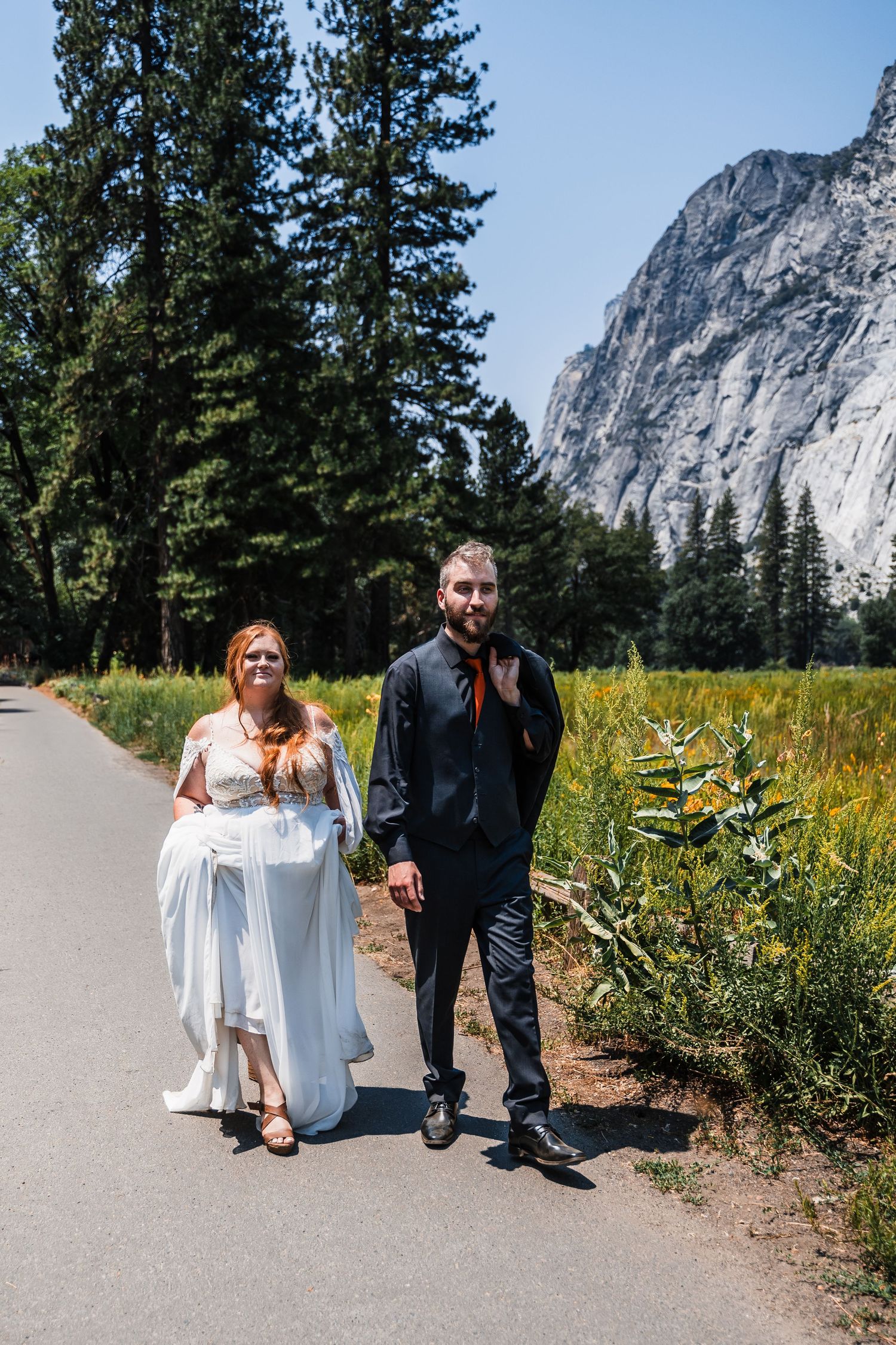 A wedding couple walk together on a paved path with the majestic granite cliffs of Yosemite Valley in the background.