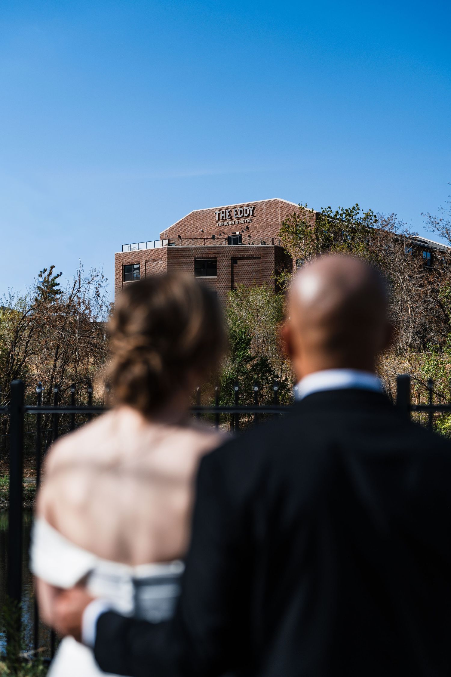 Wedding couple standing before The Eddy venue building under a clear blue sky.