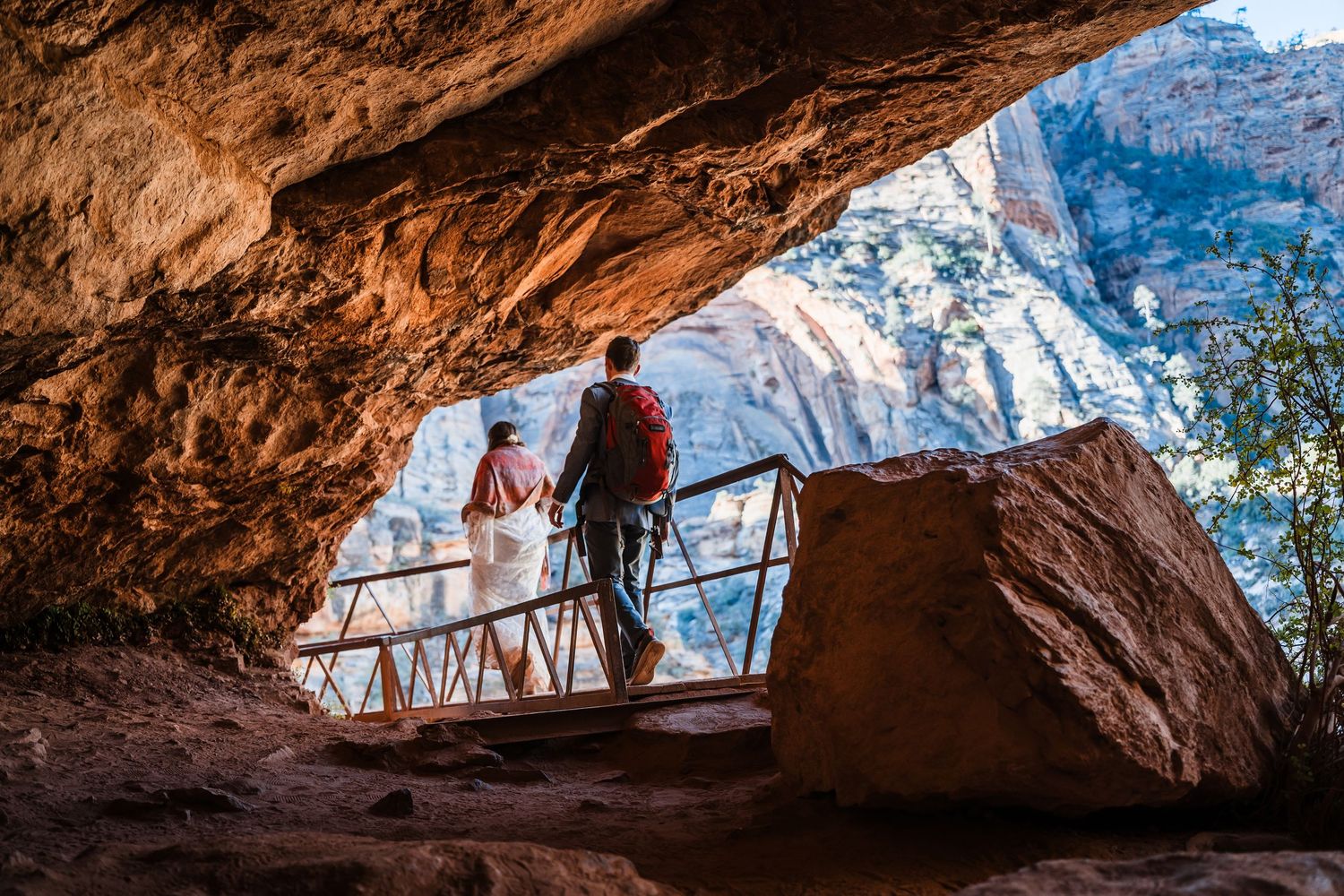 Couple standing at cave overlook viewing stunning Zion National Park canyon landscape at golden hour.