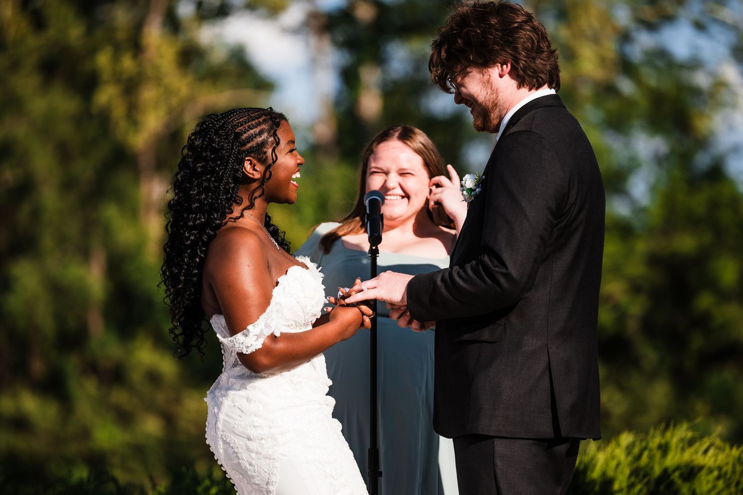 Bride and groom laughing joyfully during ring exchange at outdoor wedding ceremony.