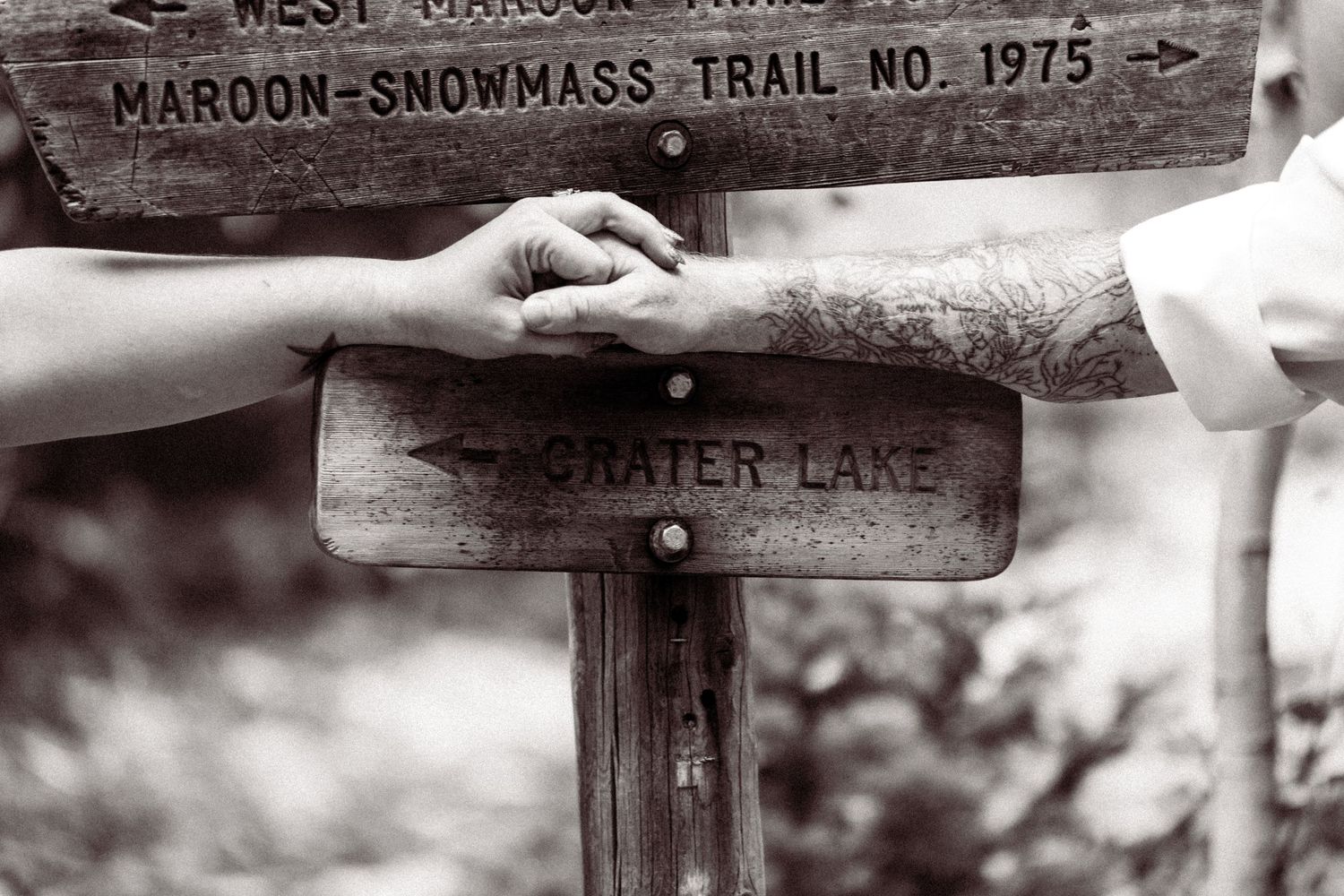 Couple holds hands near wooden trail sign for Crater Lake on Maroon-Snowmass Trail in black and white photo.