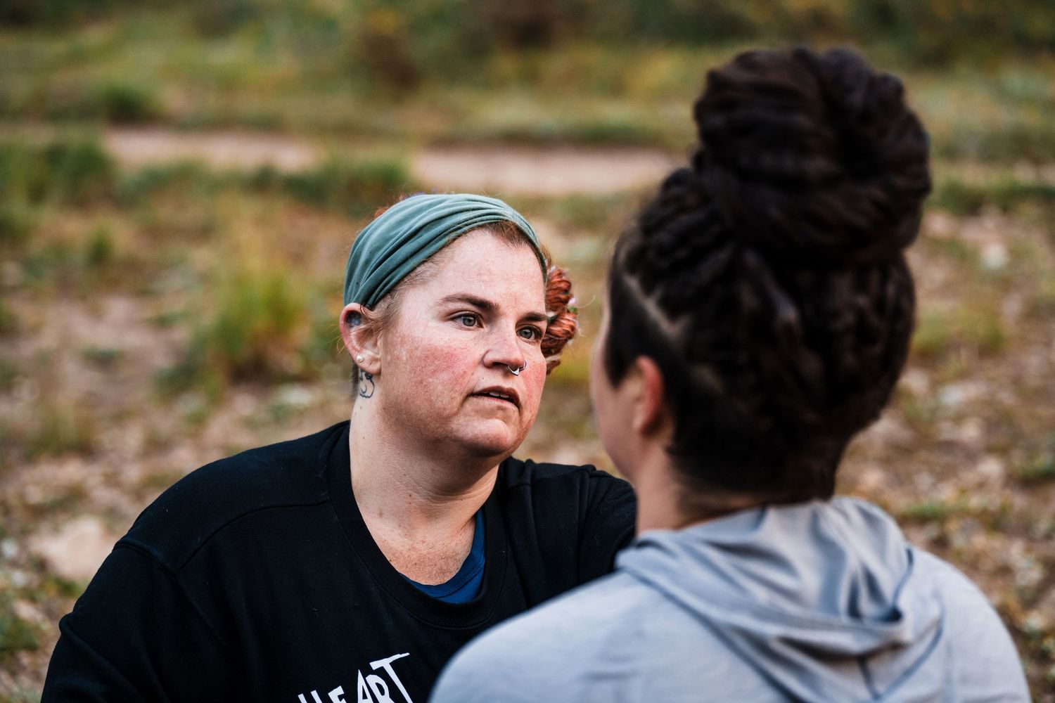 Two women having an intense conversation outdoors, one wearing a teal headband and black shirt.
