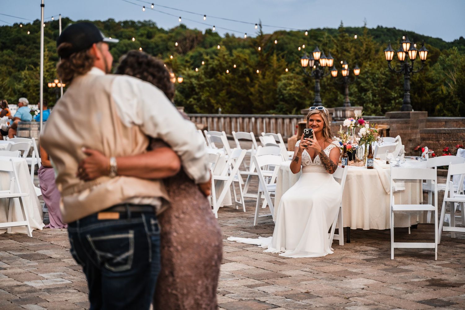 Guests mingle and dance at outdoor wedding reception with white chairs and natural lighting.