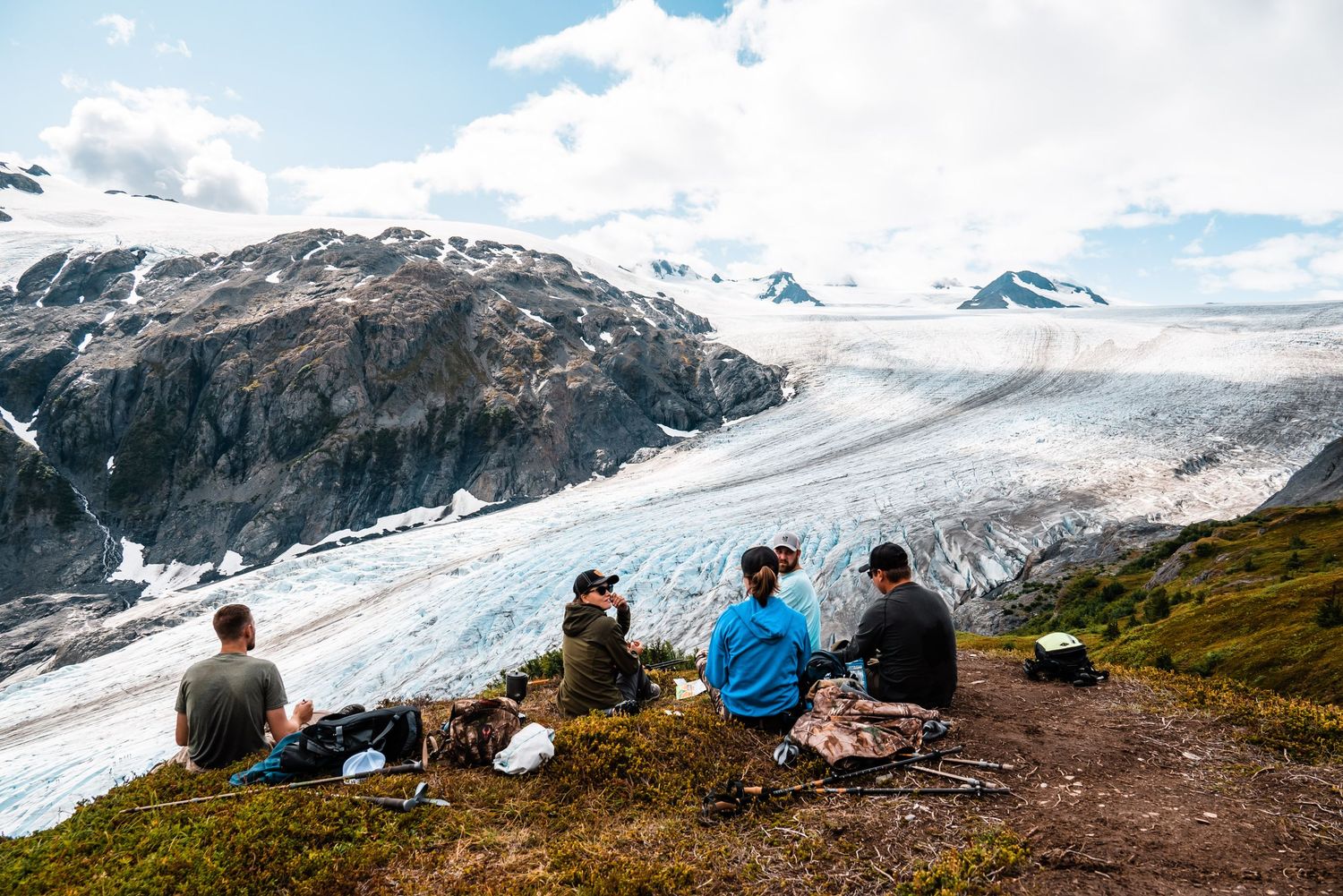 A group sits on a mountain trail overlooking a massive glacier and snow-capped peaks in a dramatic alpine landscape.