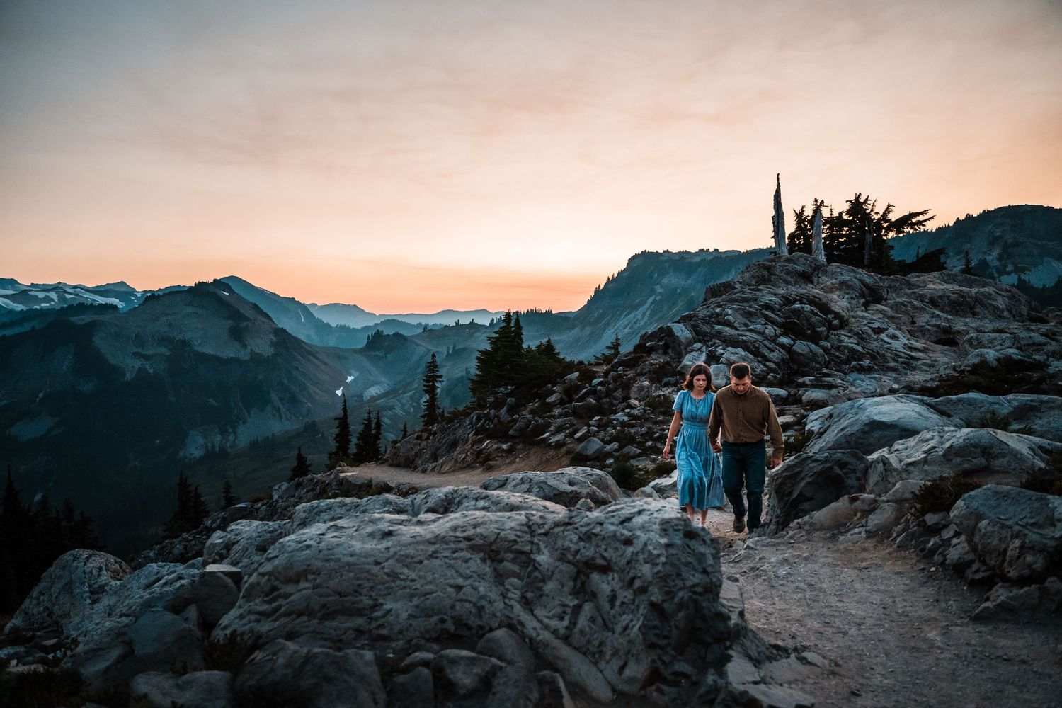 Scenic mountain landscape at sunset with rocky terrain and evergreen trees silhouetted against a pink sky.