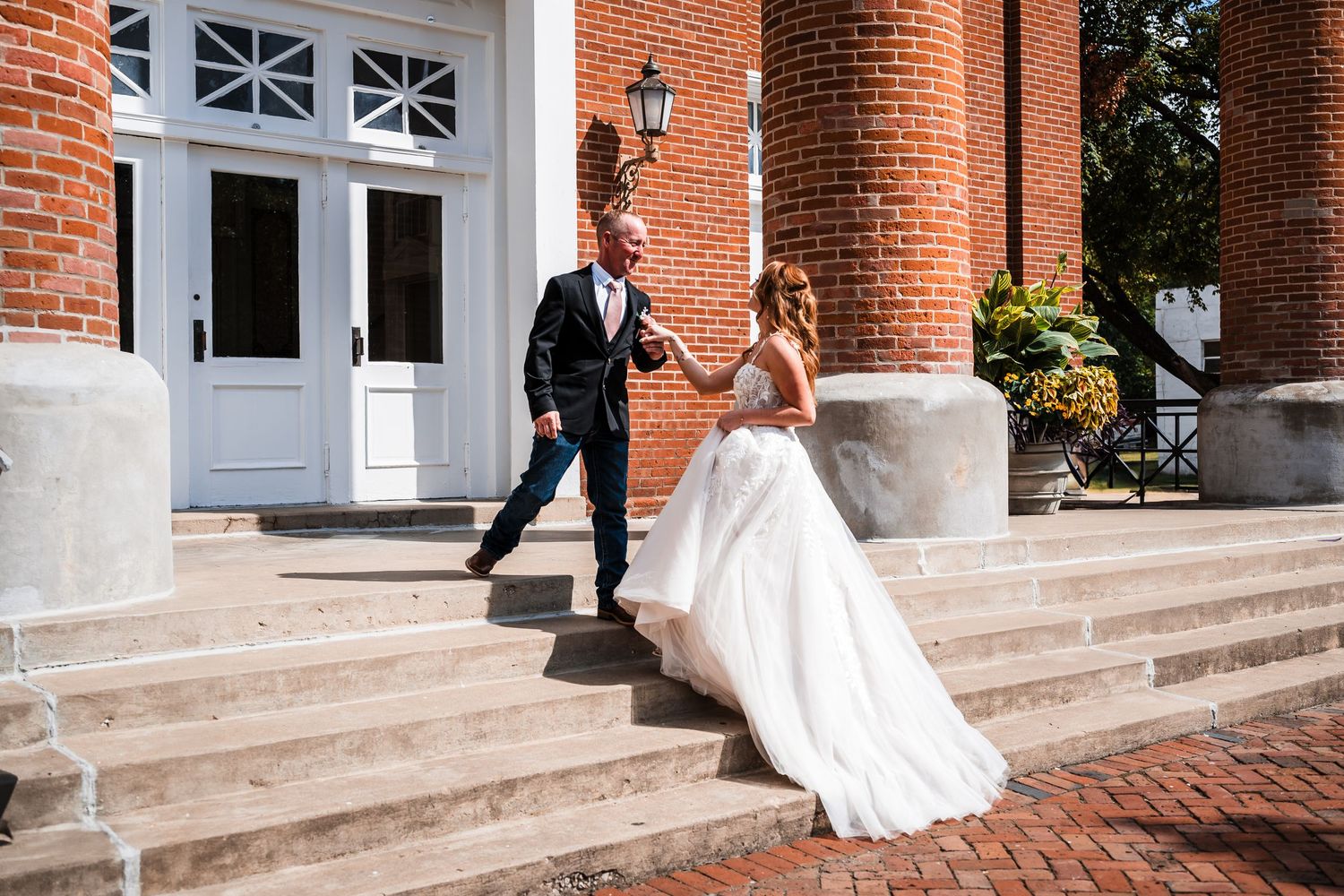 A bride in a white wedding dress walks up stone steps alongside a red brick building with white columns.