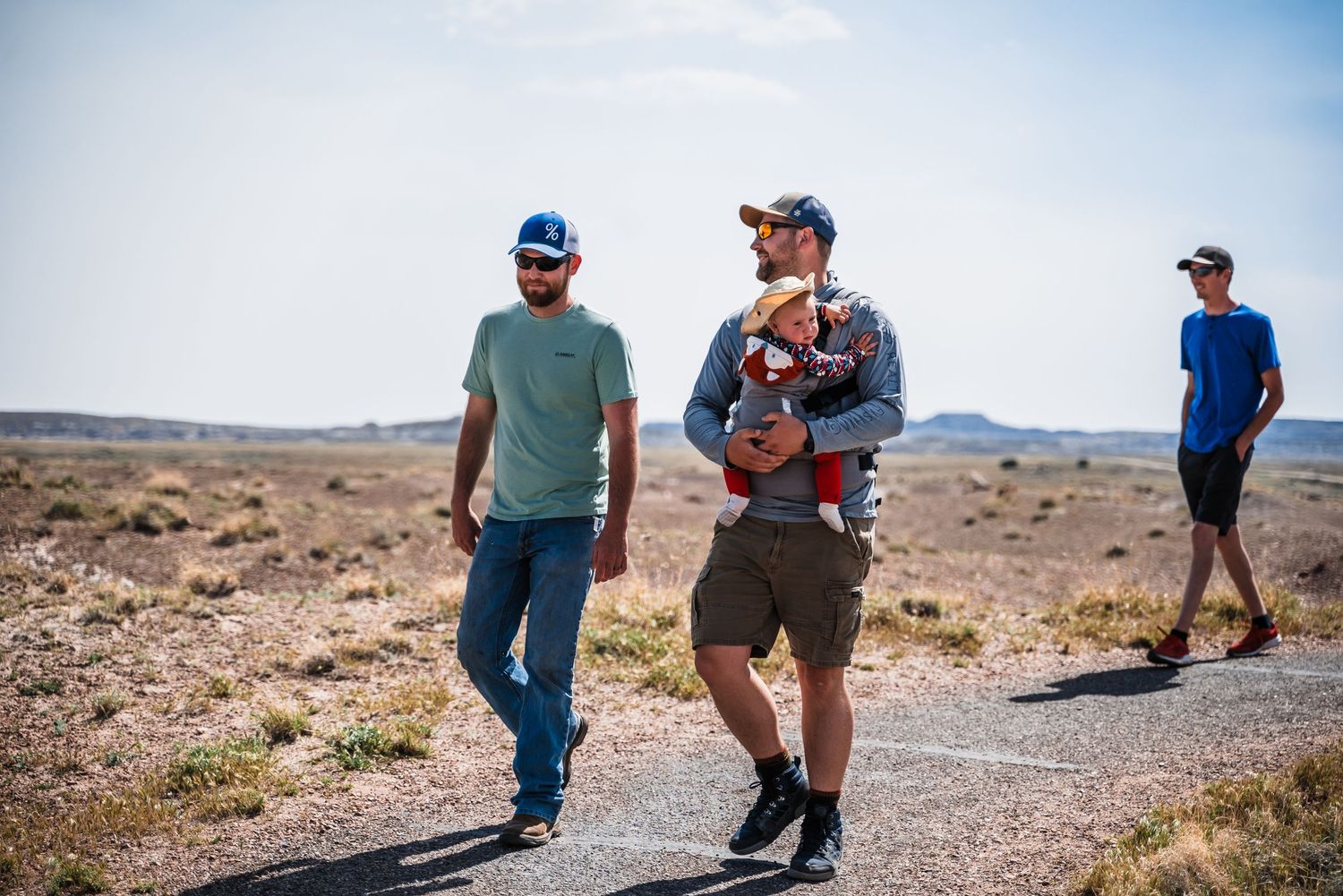 Three hikers walk along a desert trail with mountains in the background on a sunny day.