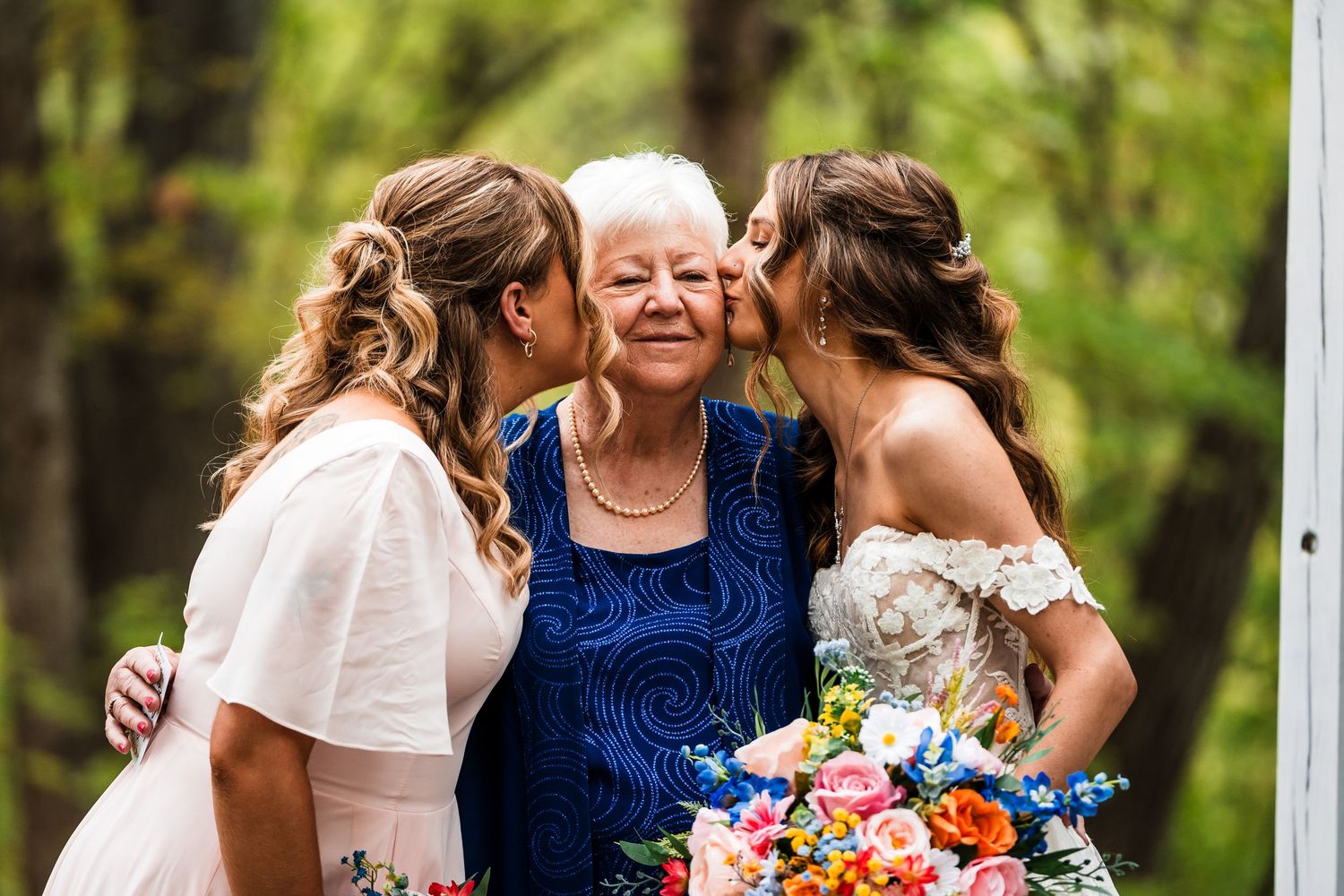 Three generations share an emotional moment during a wedding, holding colorful bouquets.