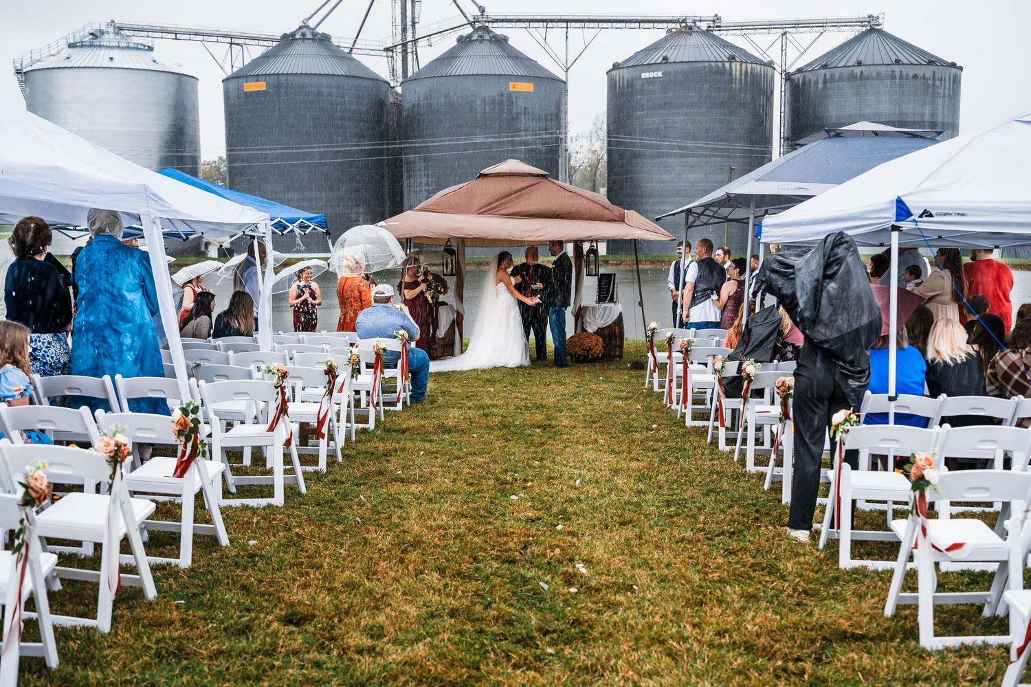 White folding chairs are set up for an outdoor wedding ceremony with grain silos visible in the background.