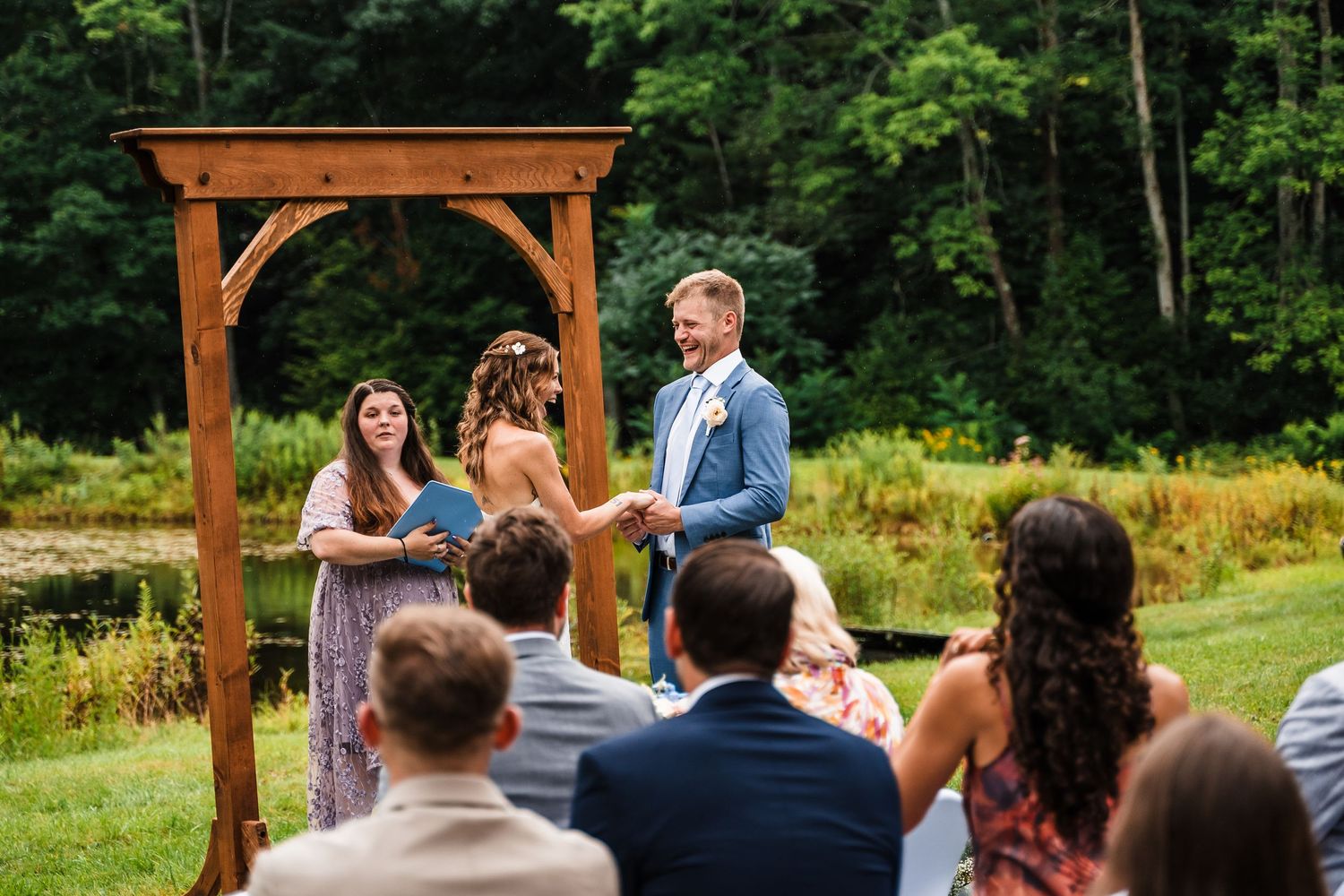 Wedding ceremony in progress under a wooden pergola with guests seated on white chairs.