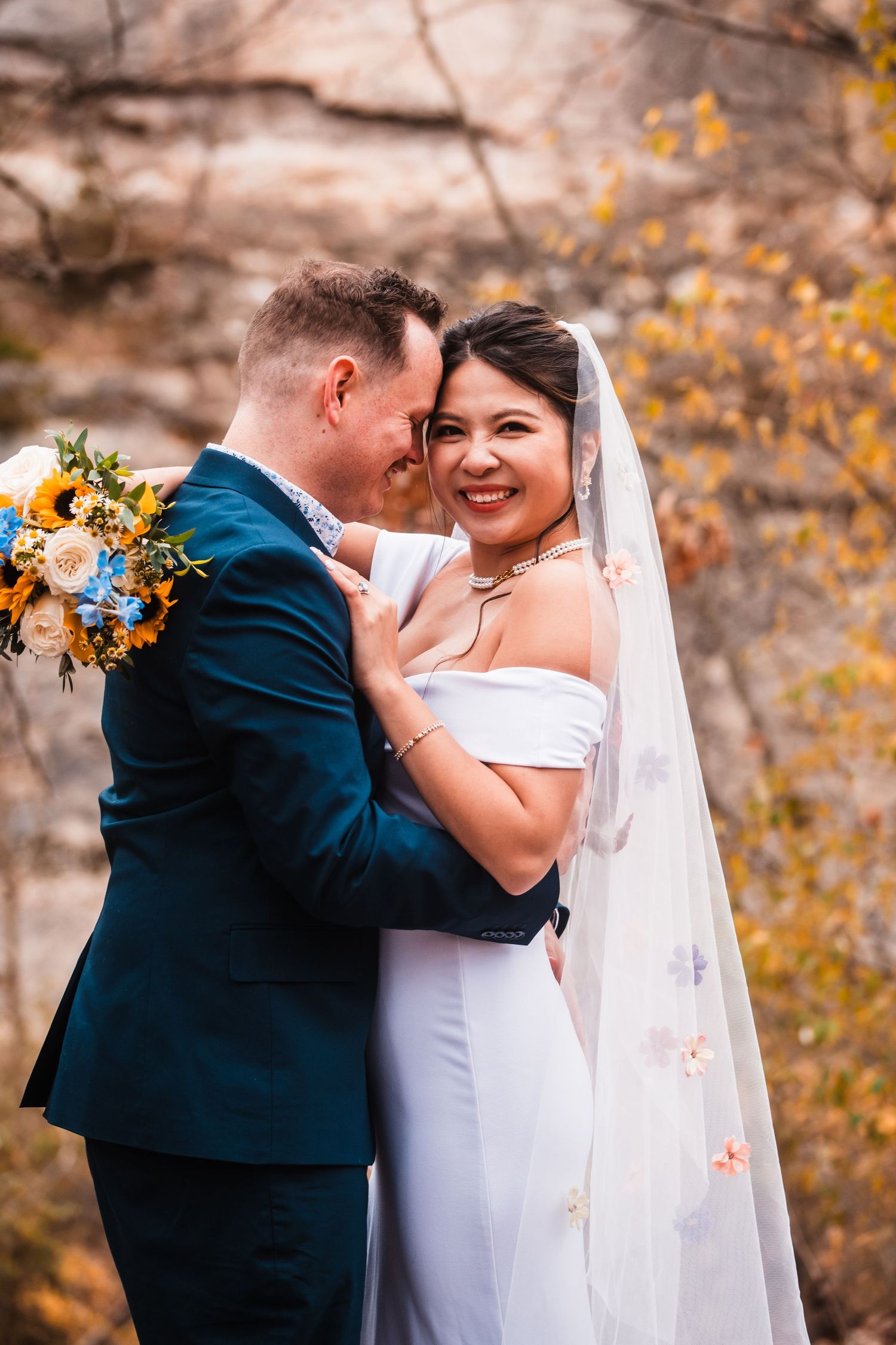 A romantic wedding portrait showing a couple embracing with autumn foliage and cliffs behind them.