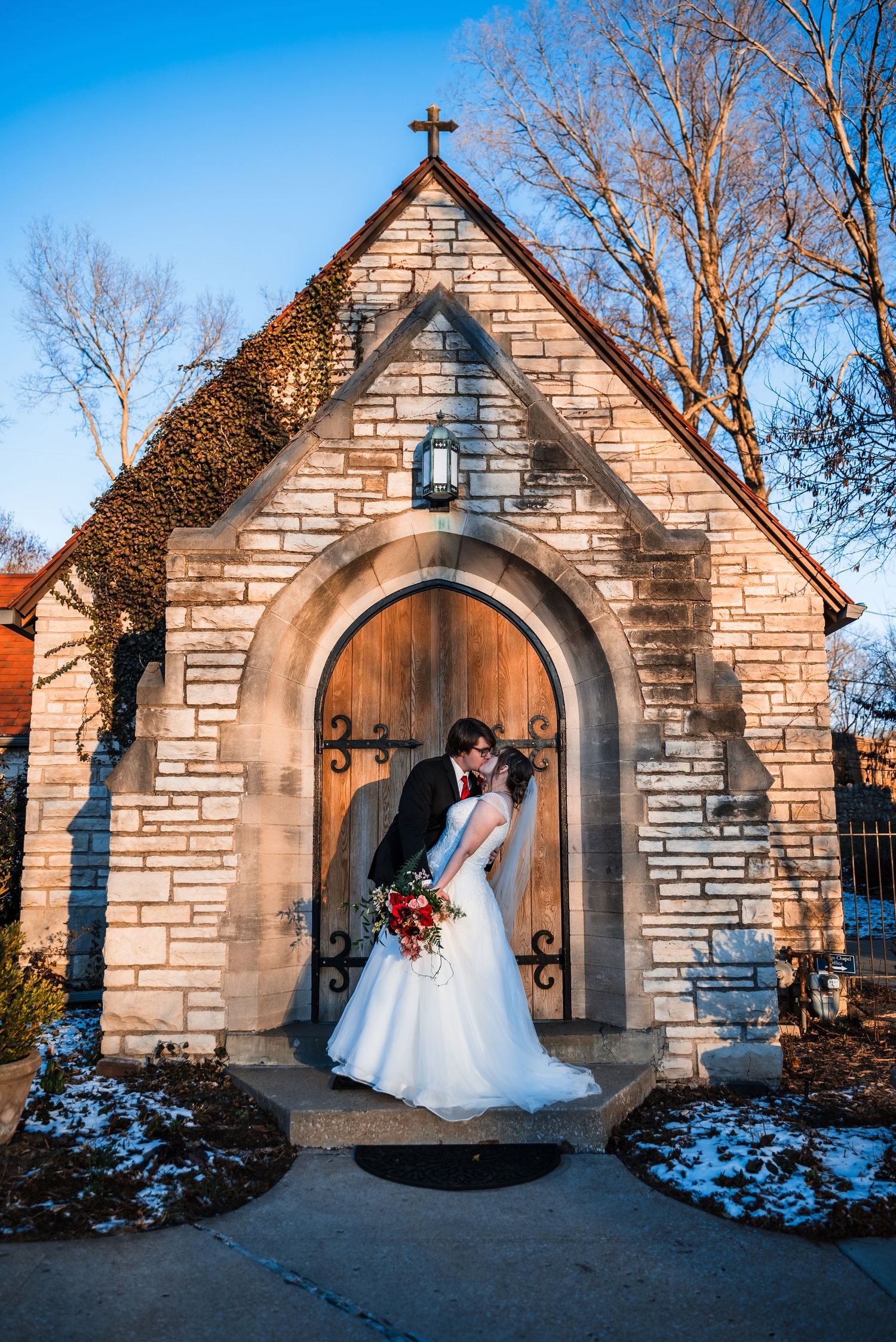 Winter wedding portrait at stone chapel entrance with snow and dramatic lighting.