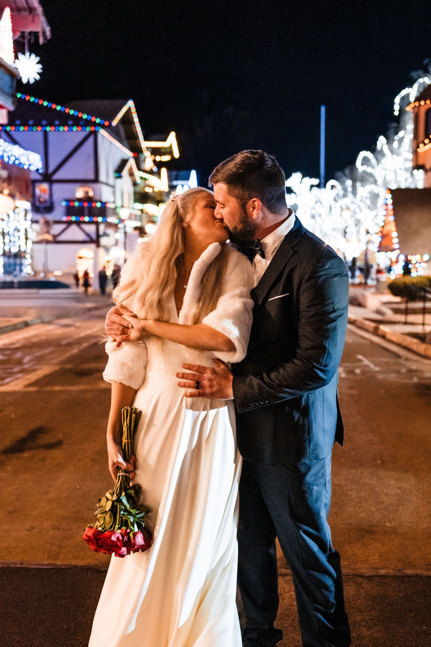 A romantic wedding moment on a festive street with twinkling lights and holiday decorations.