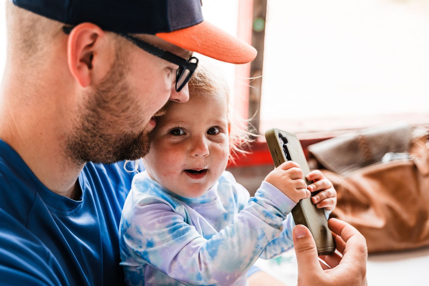 A child in tie-dye shirt looks at a mobile device with an adult wearing a cap and glasses.