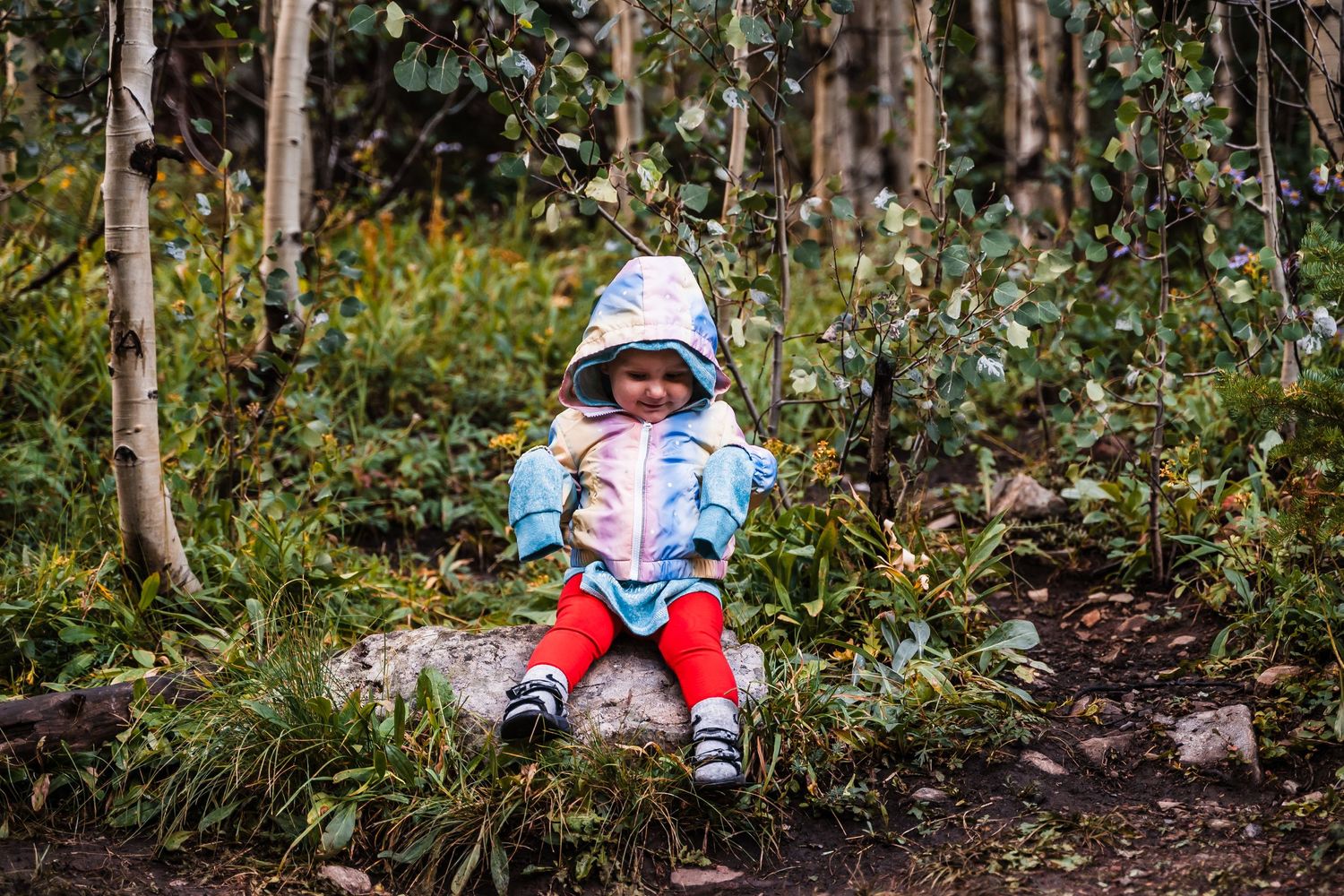 A small child sits on a log in a forest wearing winter clothing and a sun hat.