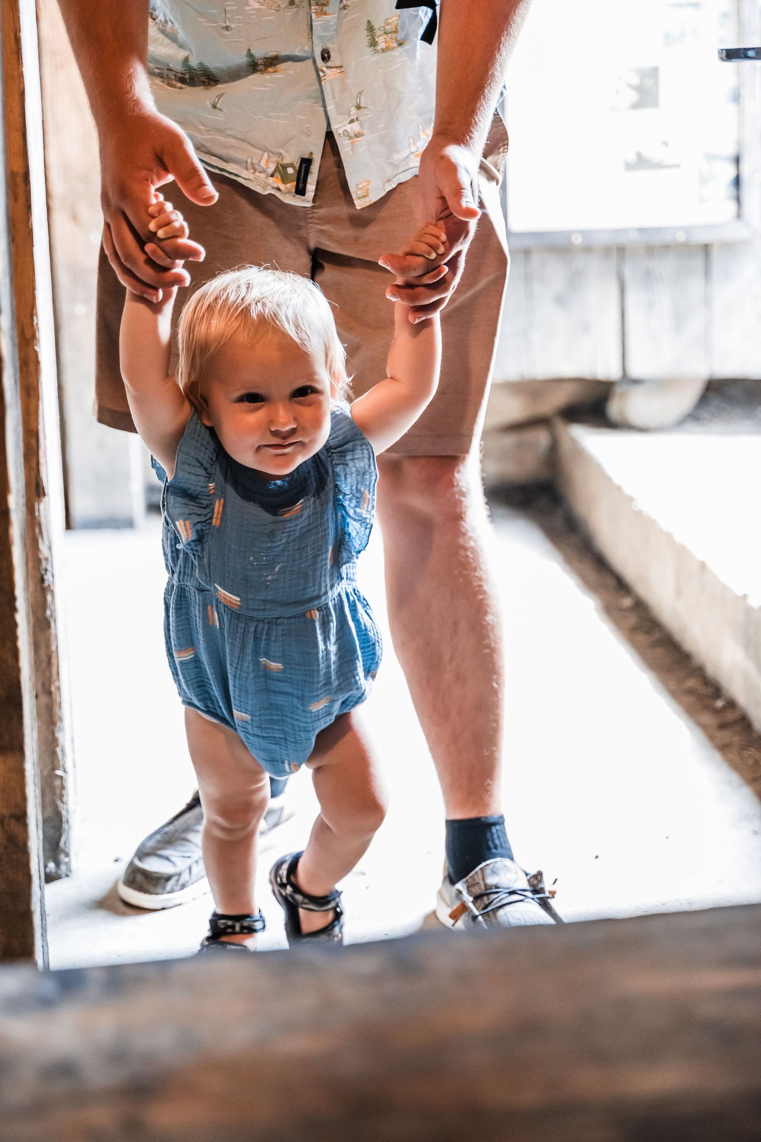A young child wearing blue denim overalls takes first steps while holding adult's hands for support.