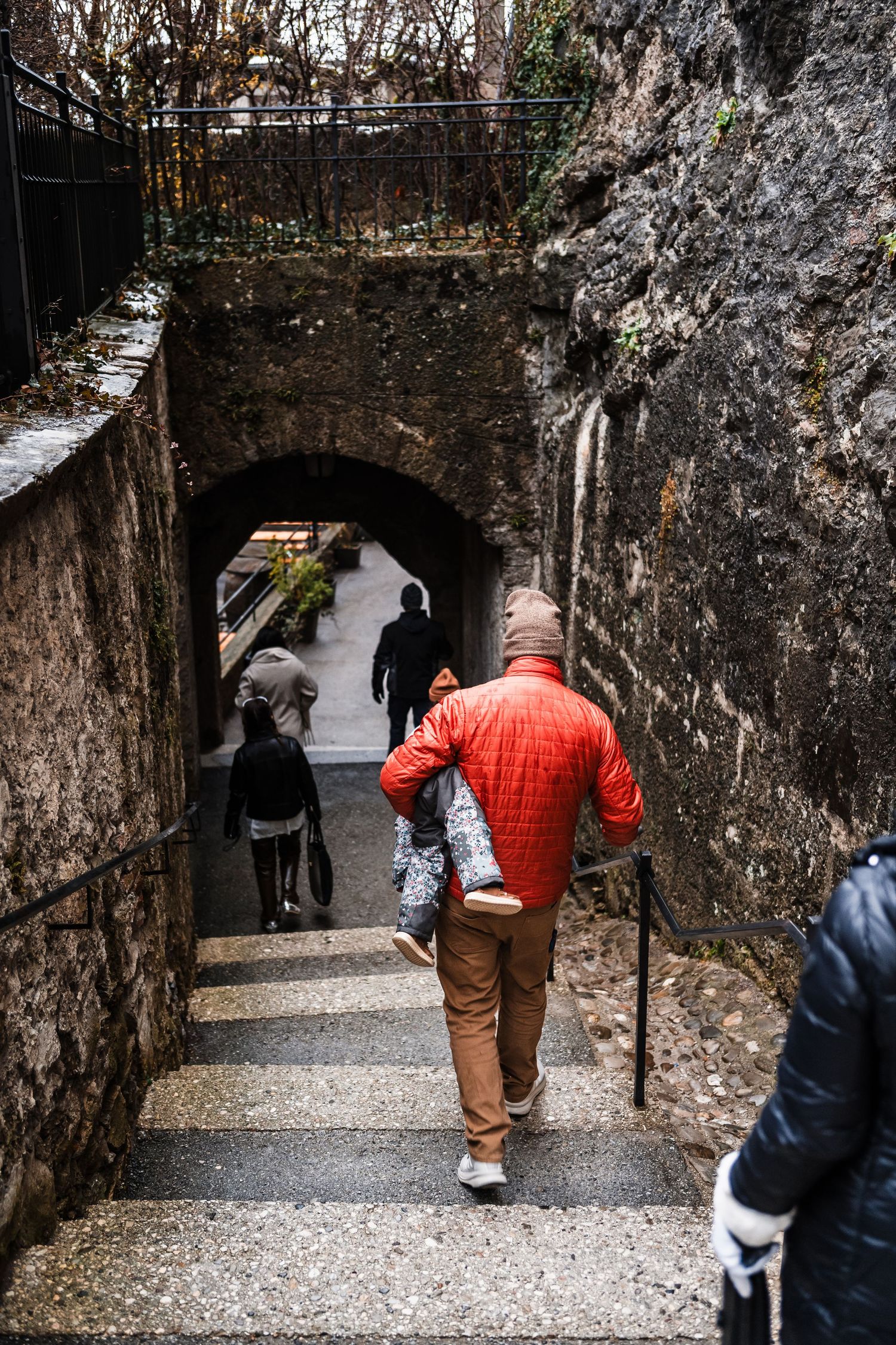 People walking down stone-walled pathway with archways in historic European setting.
