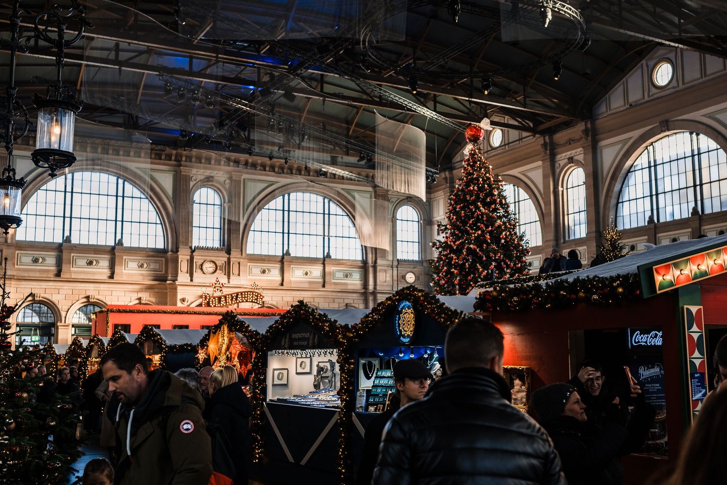 Panoramic view of a historic indoor marketplace with arched windows, Christmas trees, and festive holiday decorations.