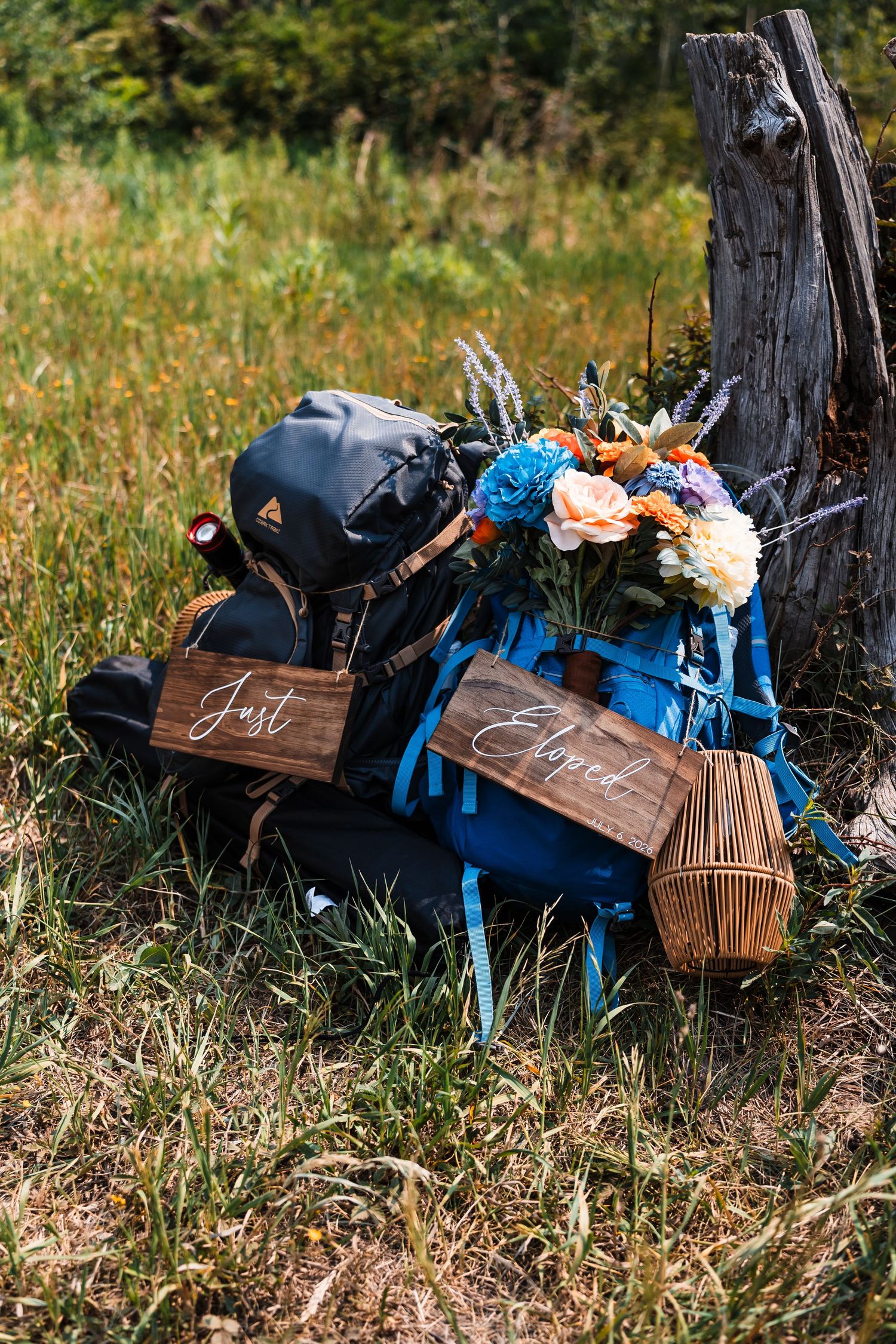 Just Eloped wooden signs rest against hiking backpacks with colorful wildflower bouquet outdoors.
