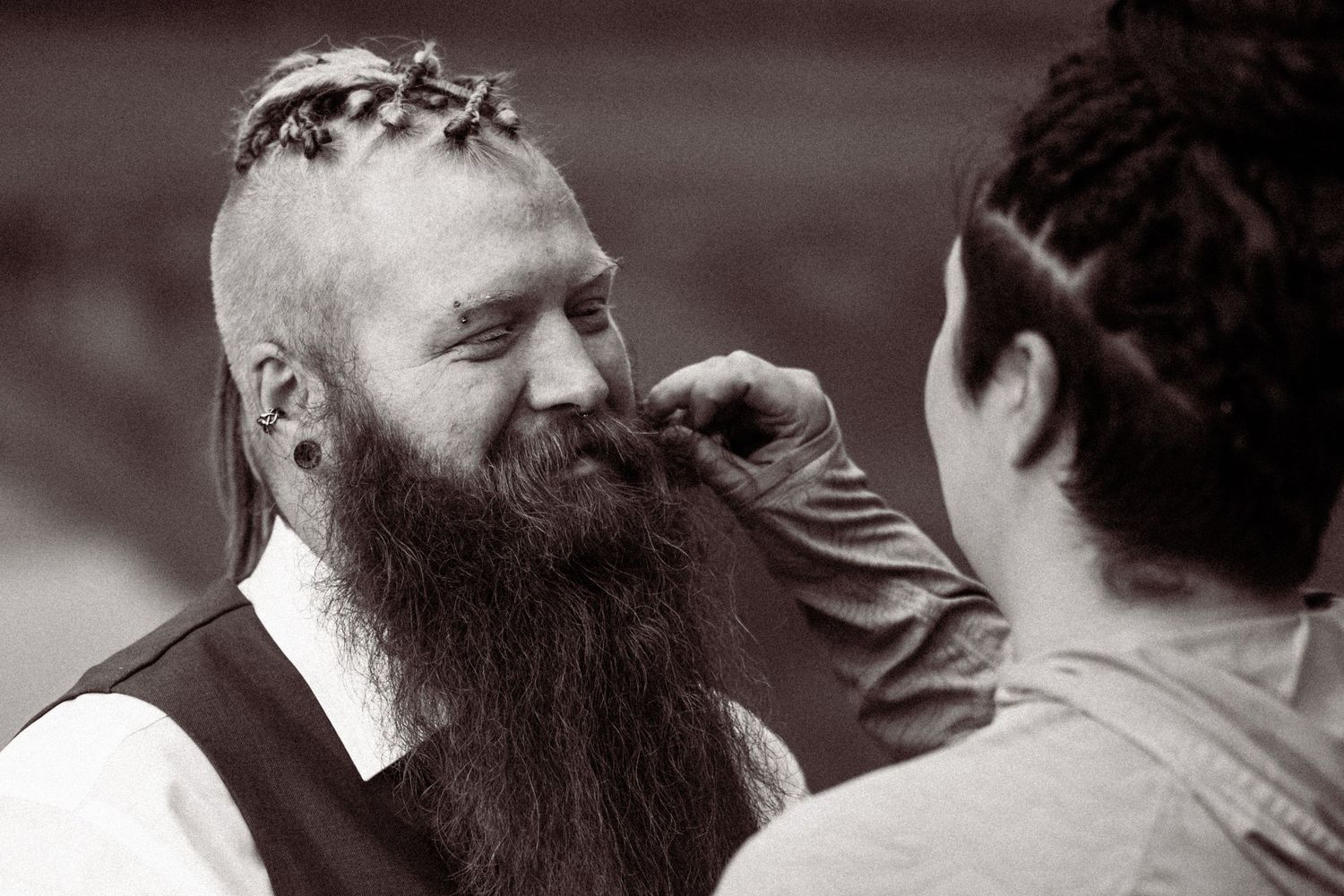A bearded man with a braided mohawk has his beard groomed by another person in a black and white photo.