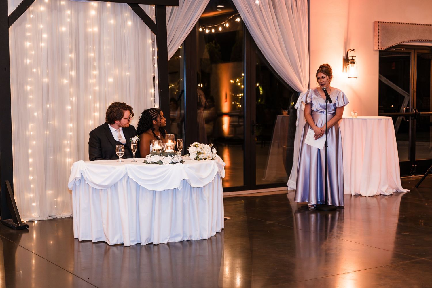 Bride and groom at sweetheart table while bridesmaid delivers speech at elegant reception.
