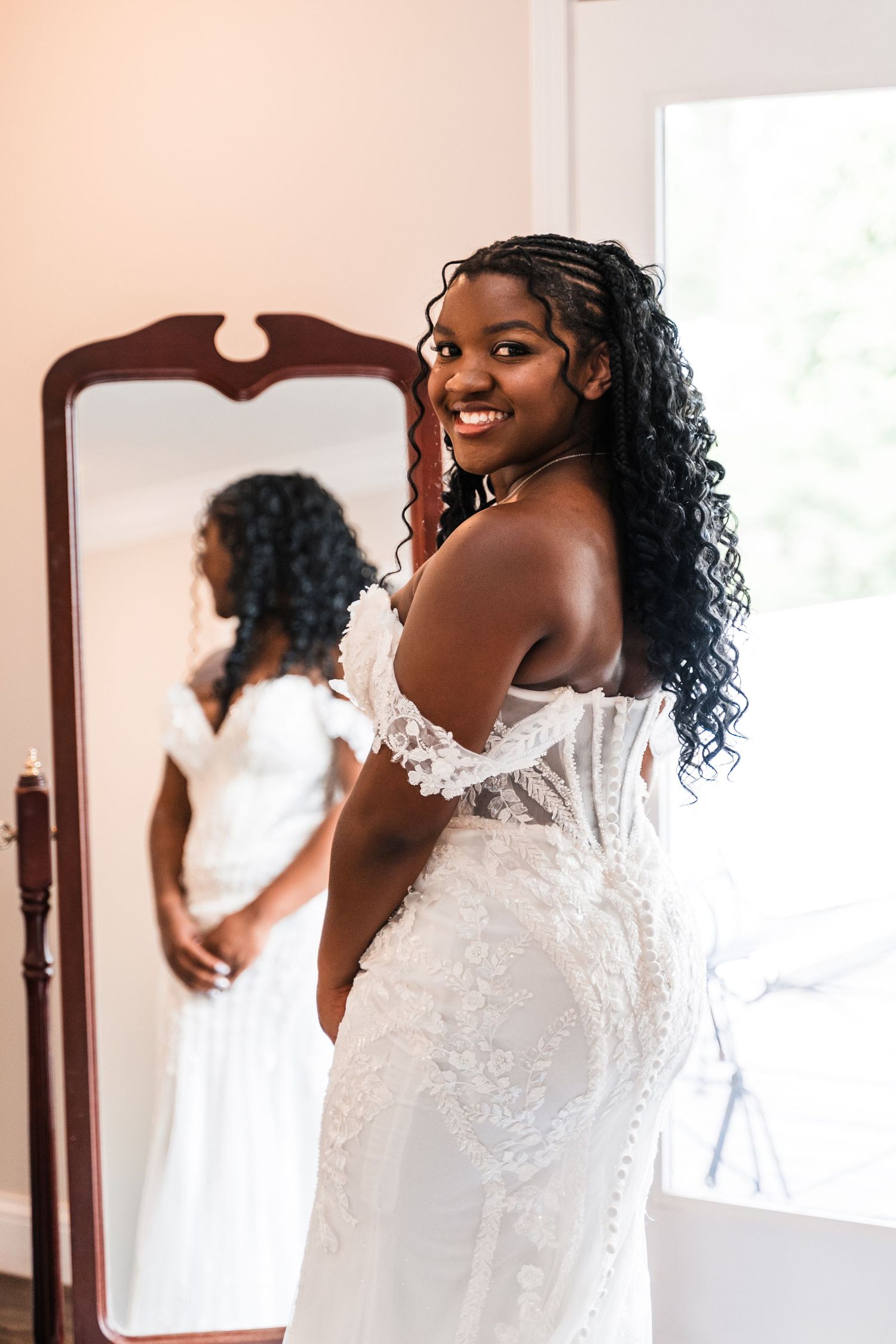 Smiling Black bride in elegant lace wedding gown posing in front of full-length mirror.