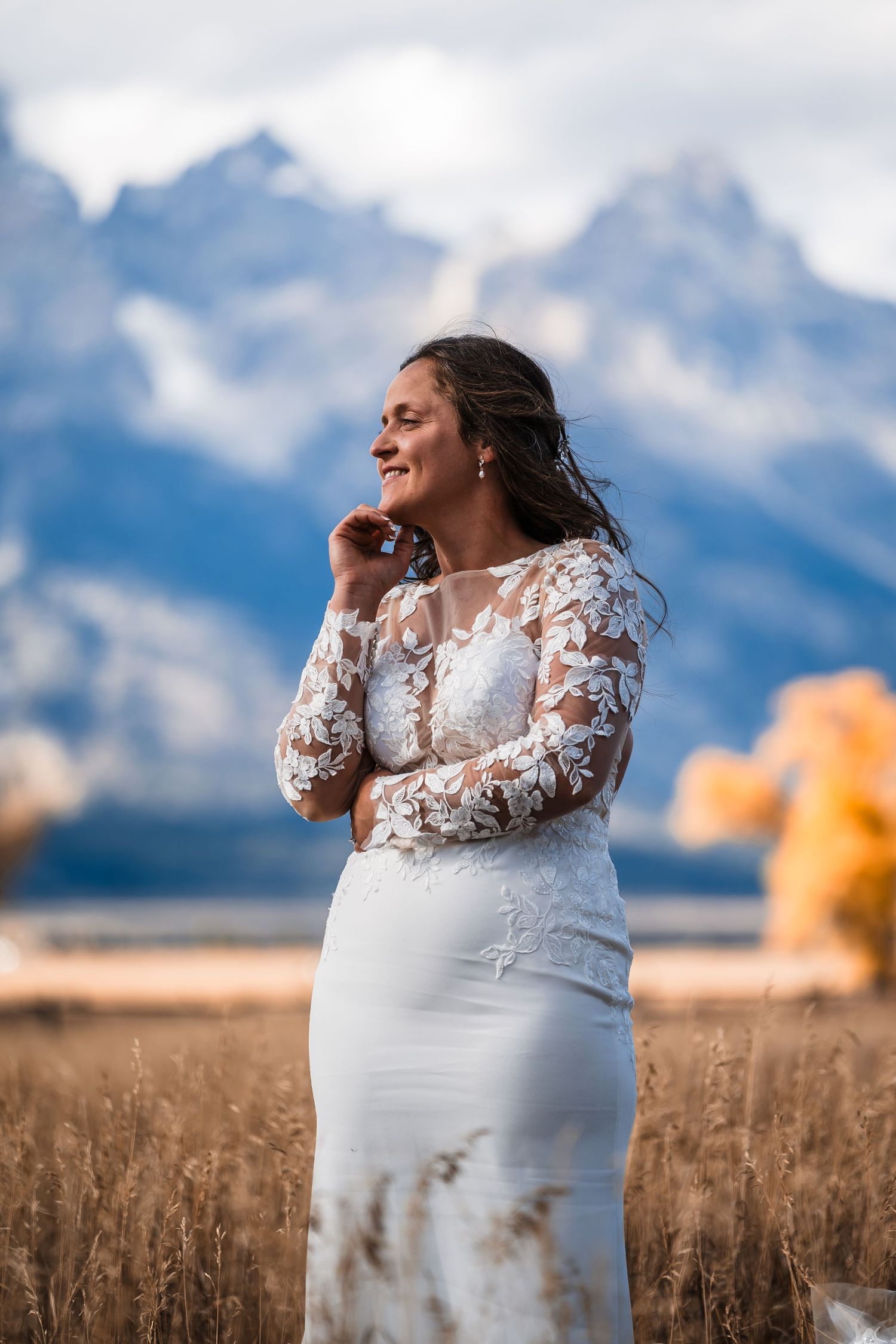 A bride in lace stands in a golden field with majestic mountain peaks rising in the misty background.