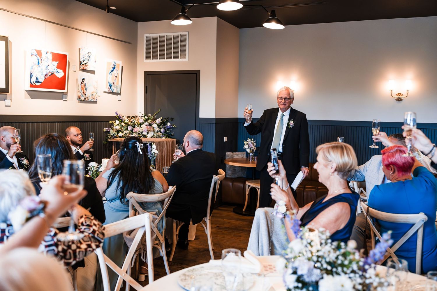 Elder guest raises glass for toast during wedding reception in elegant art-filled dining room.