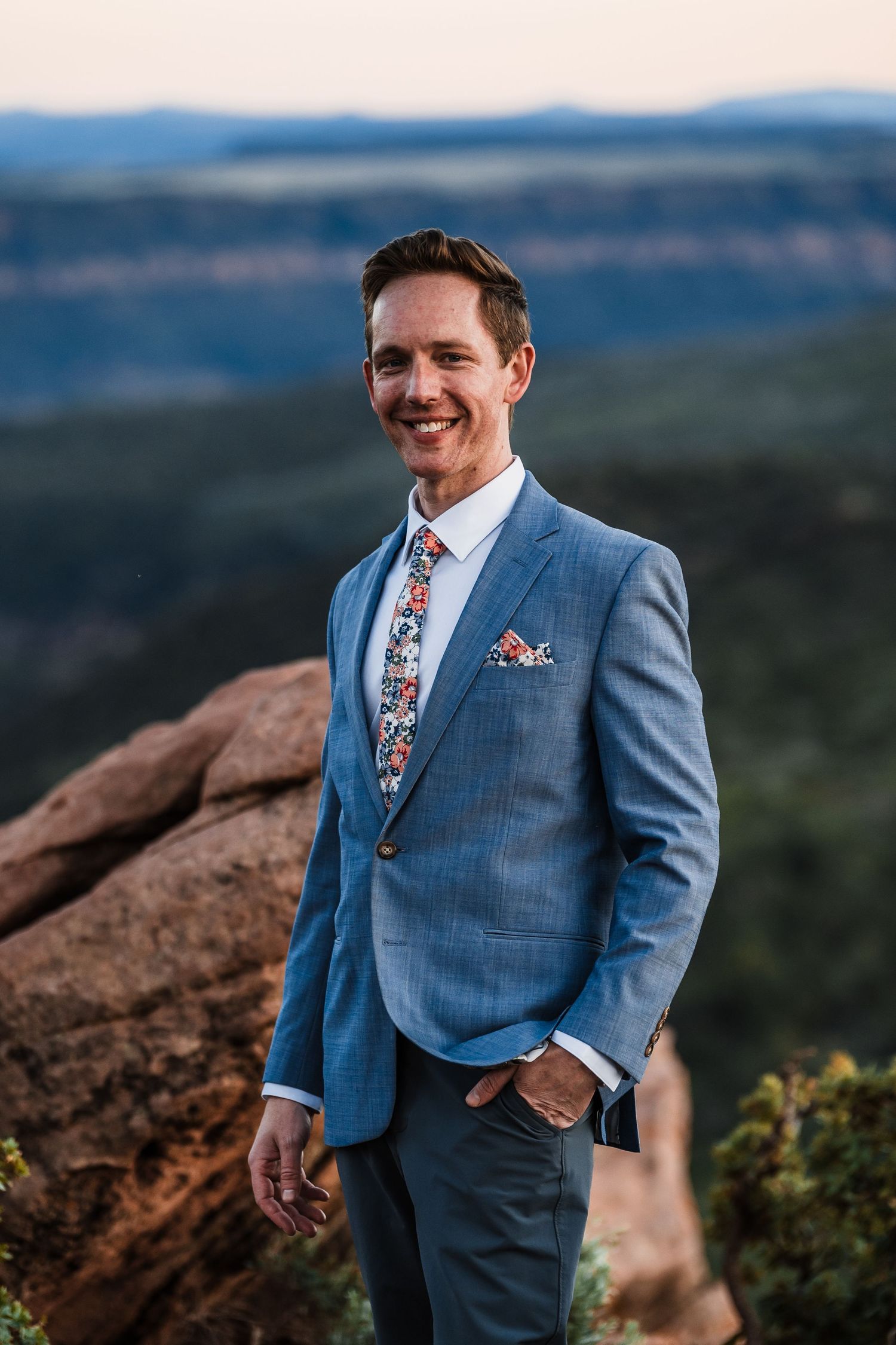 Smiling groom in light blue suit with floral tie poses on red rock overlook at sunset.