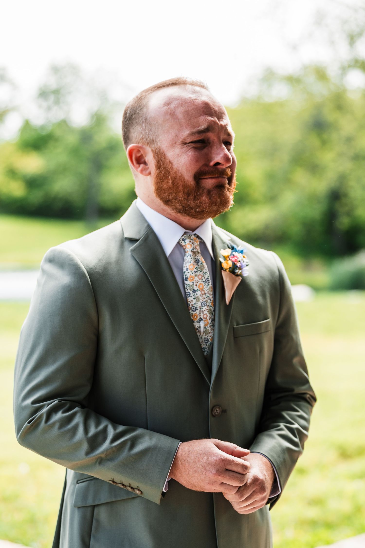 Emotional groom in sage green suit with floral tie and boutonniere tearing up outdoors on wedding day.
