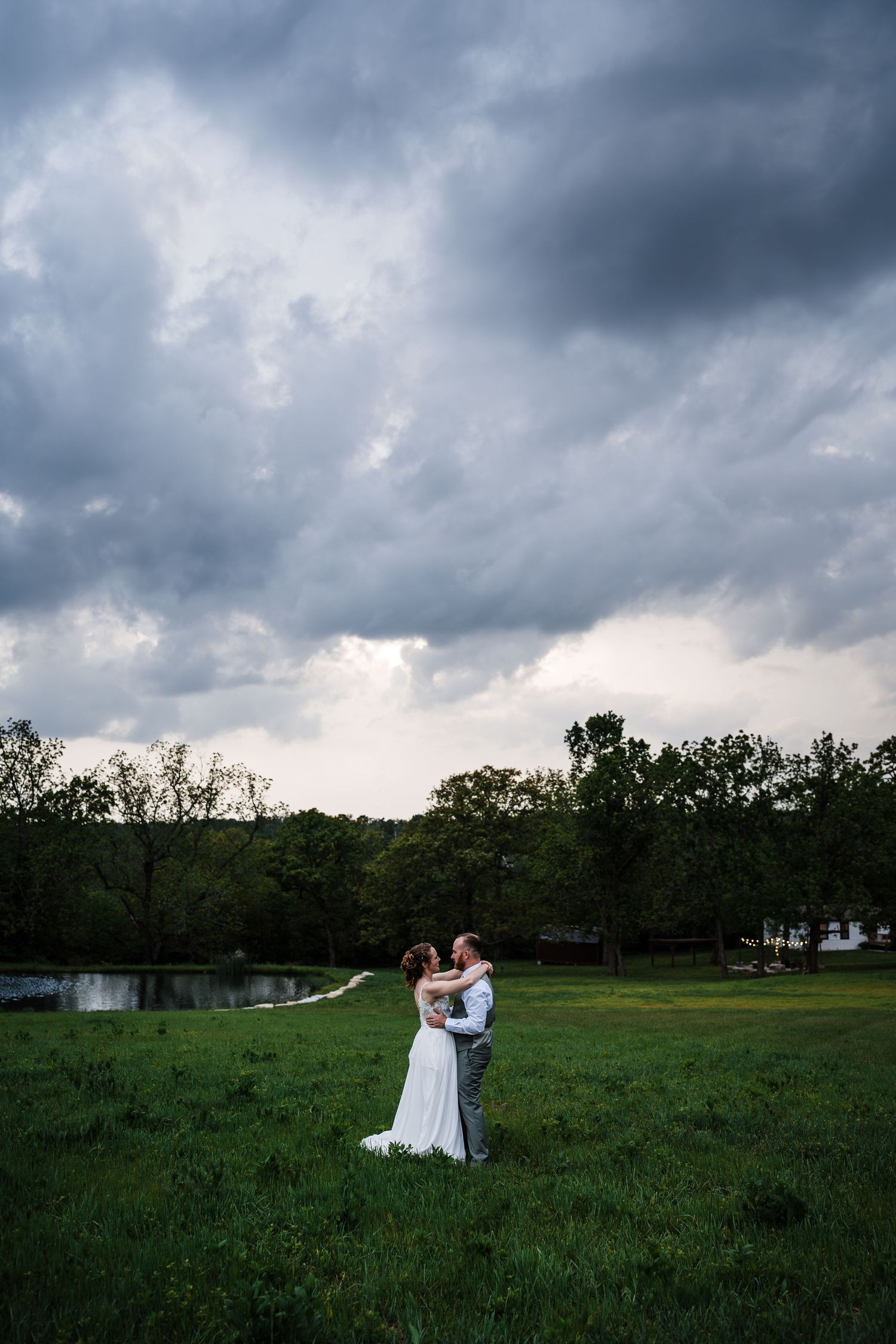 Bride and groom embrace in green field under dramatic stormy sky at golden hour.