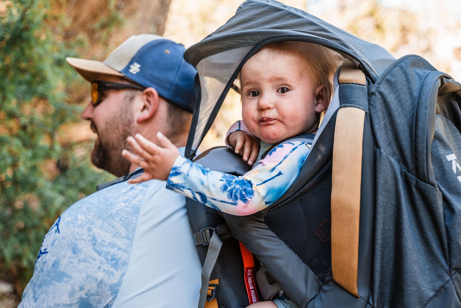 A baby peeks out from a child carrier backpack while hiking outdoors on an autumn day.