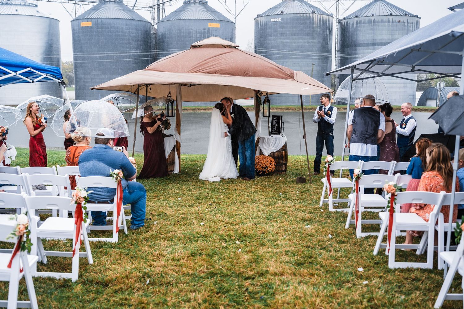 Outdoor wedding ceremony setup with white folding chairs and grain silos visible in the background.