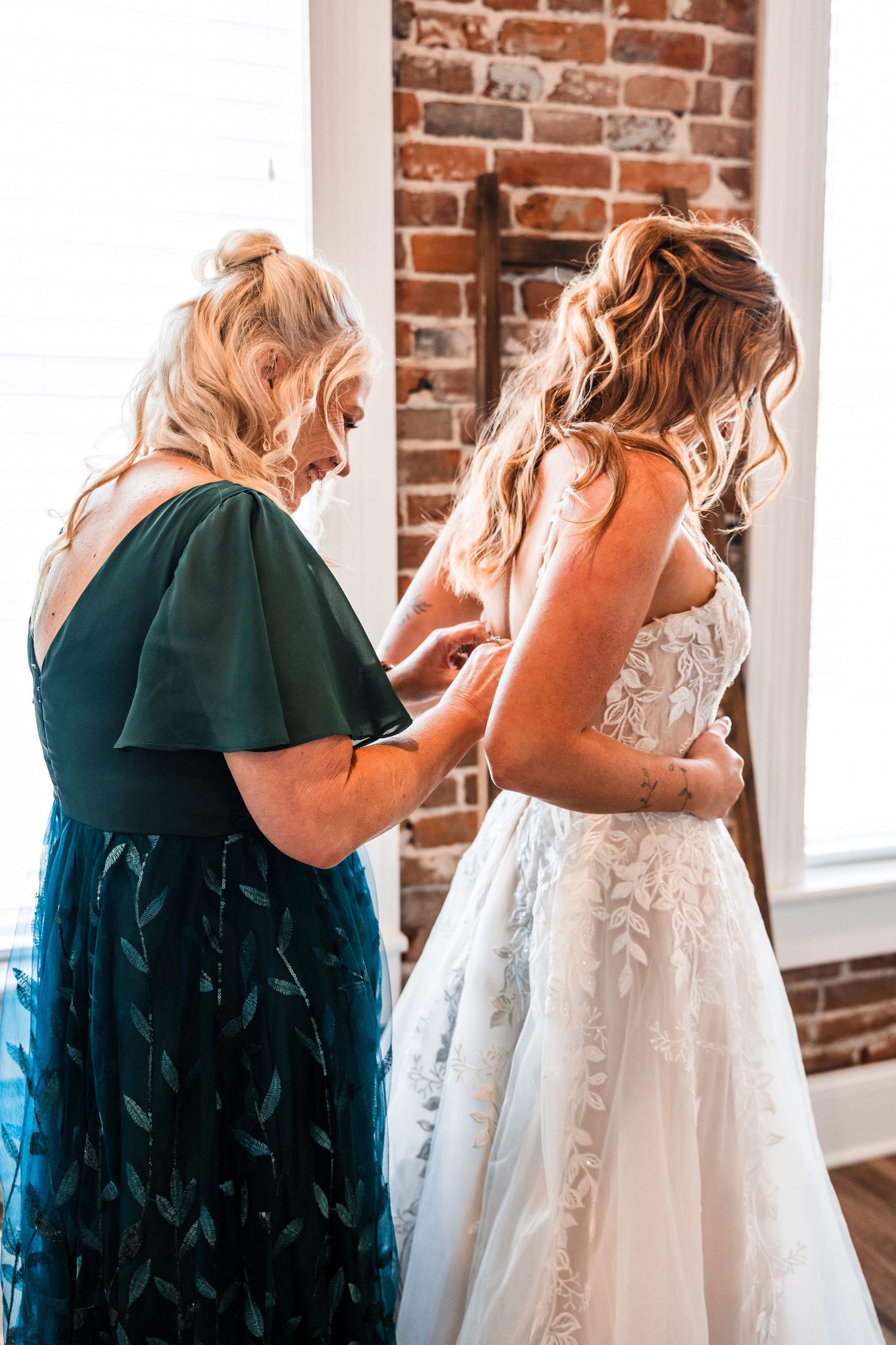 Bridesmaids in dark teal dresses help the bride prepare by buttoning up her ornate white wedding gown.