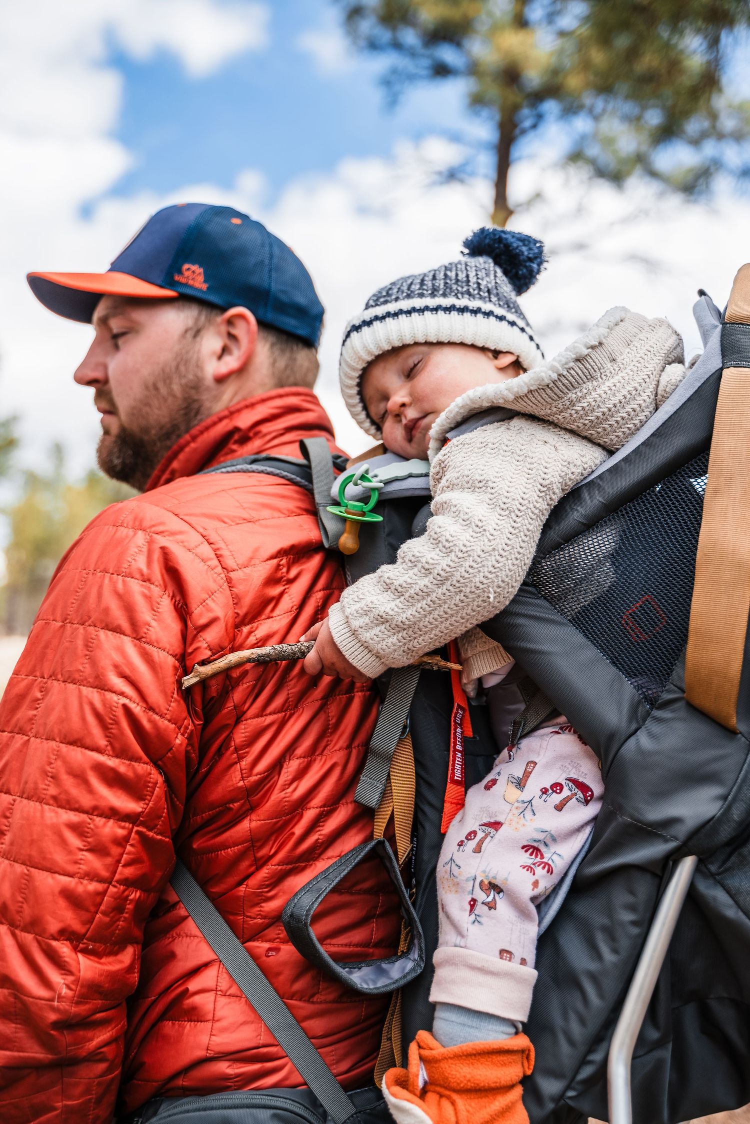 A baby sleeps soundly in a hiking carrier backpack while being carried through a sunny forest.