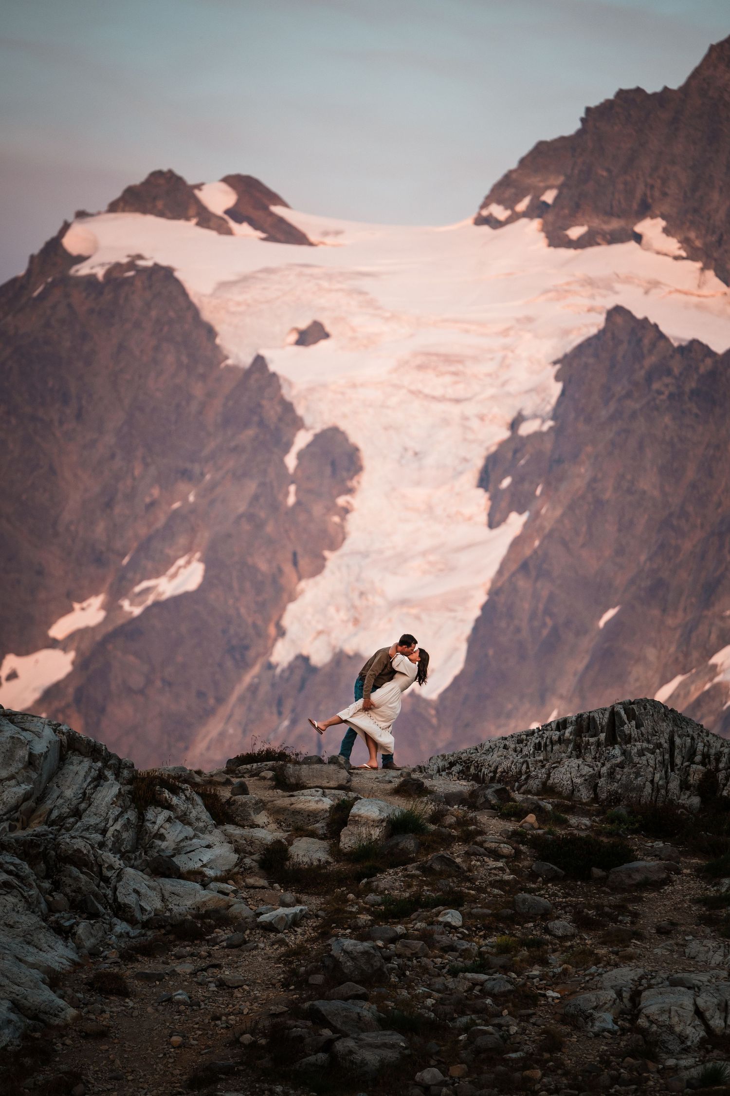 Silhouetted figures stand on rocky mountain ridge with snow-capped peaks glowing in warm sunset light.