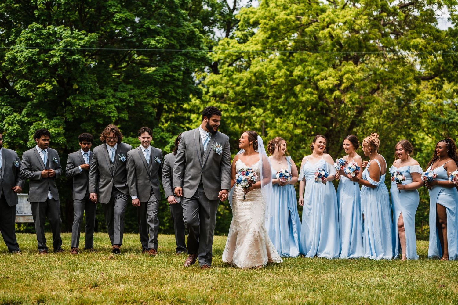 Wedding party in blue and grey attire posing together on a lush green lawn.